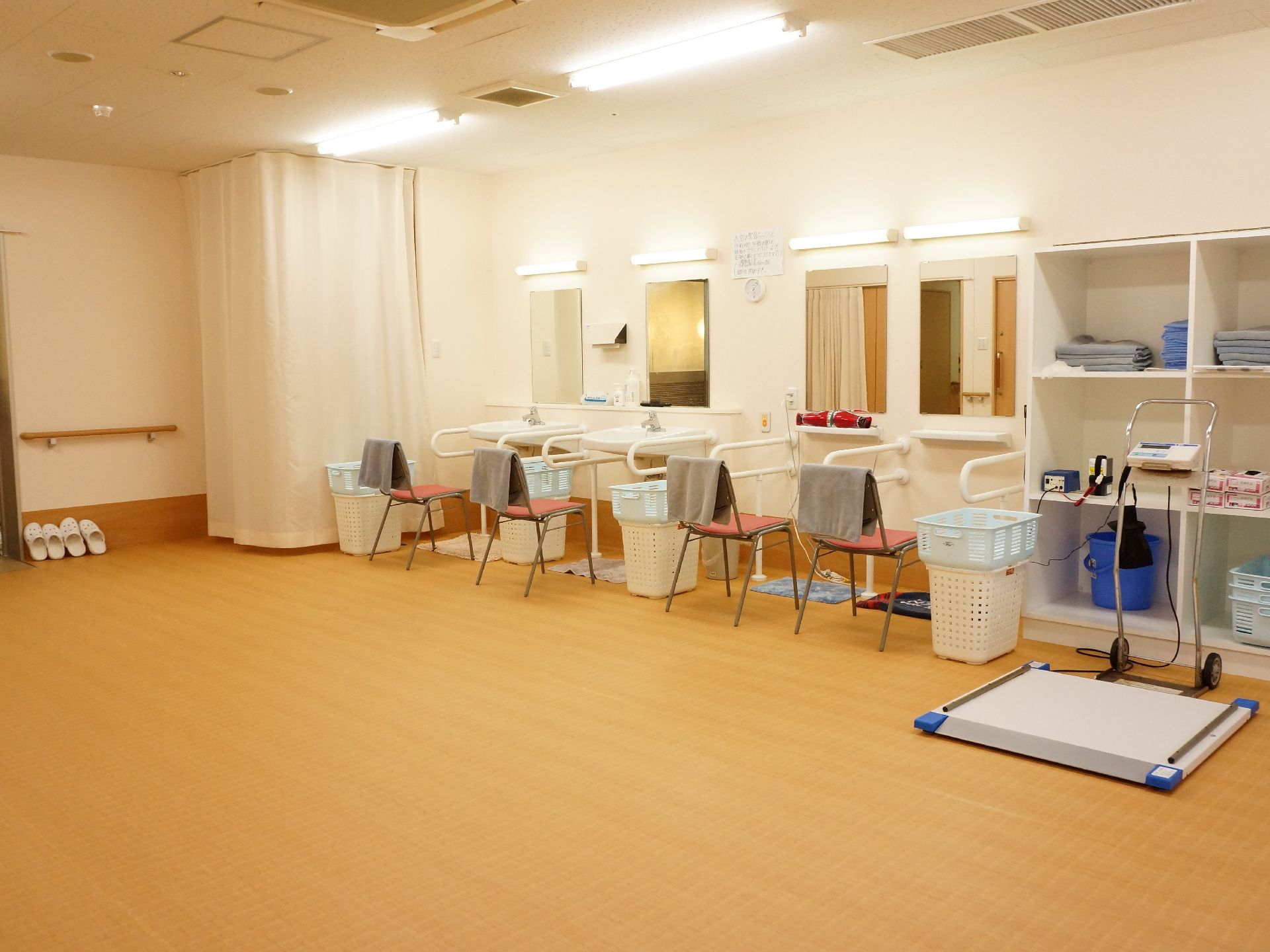 脱衣室には、体重計が置かれている。部屋の右手に棚があり、タオル類やカゴがある。カーテンで仕切られたスペースもある。