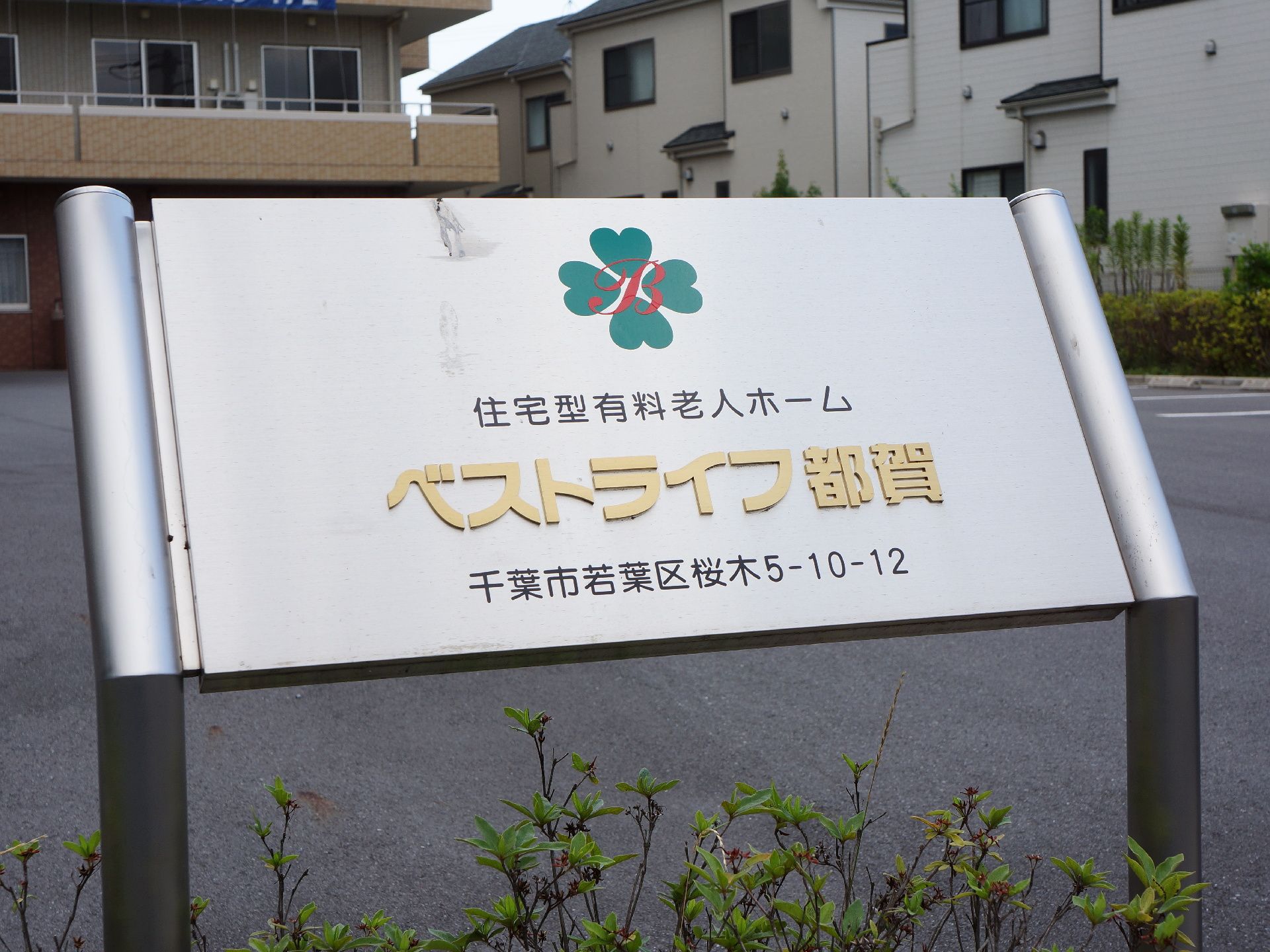 施設の敷地入口に、看板が設置されている。鉄の板に、施設名と住所が記載されている。看板の下には、苗木が植えられている。