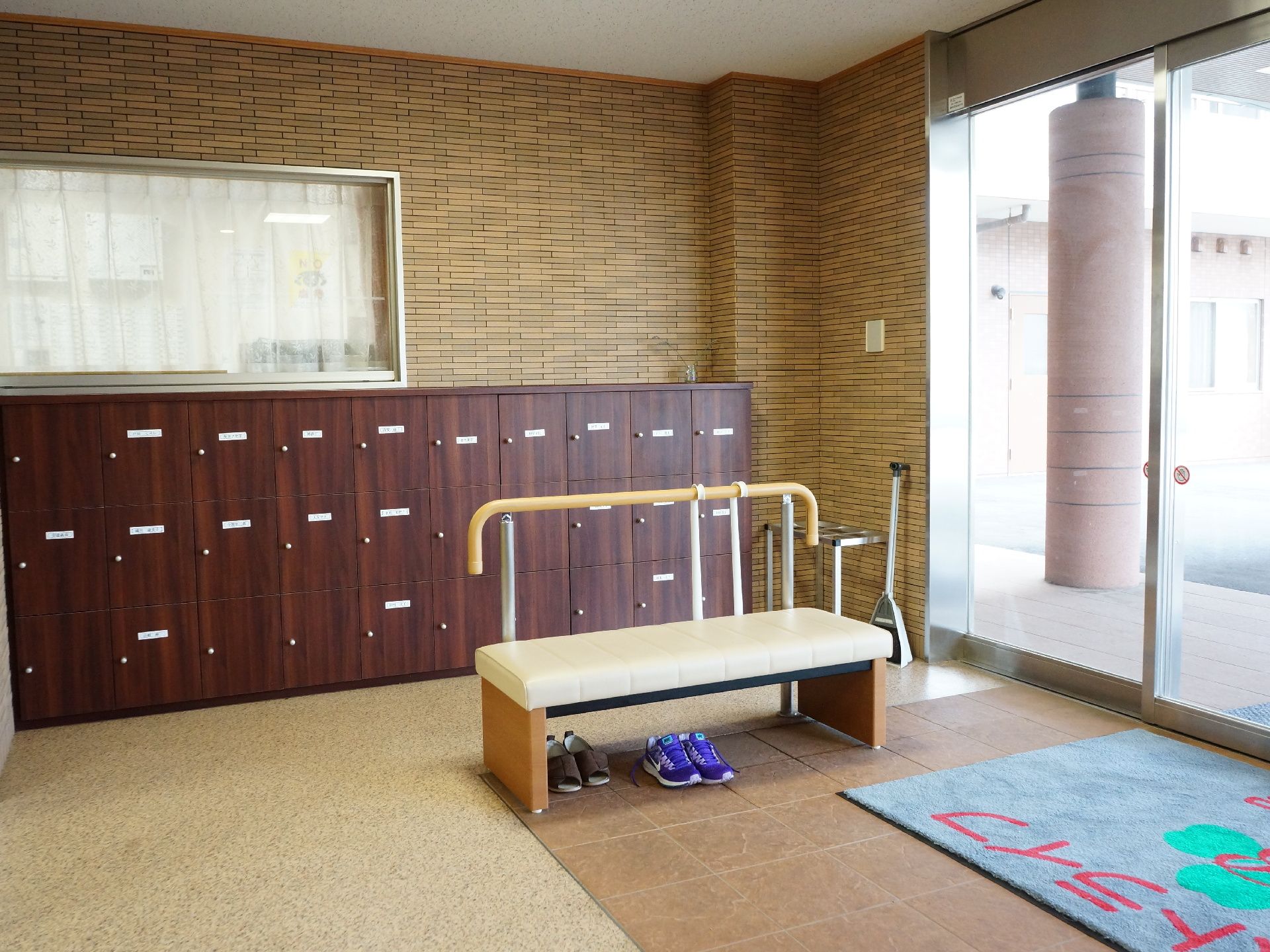 施設の玄関右手には、手すりが設置されており、その前に横長の椅子が置かれている。右奥には、下駄箱が並んでいる。