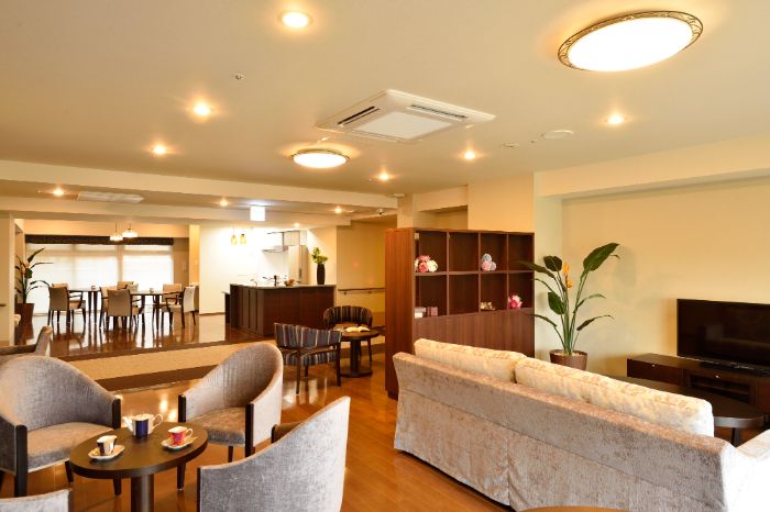 施設のラウンジは白の天井とグレーの壁、フローリングの床。奥には窓があり中央にテーブルと椅子が置かれている。トイレなども見える。