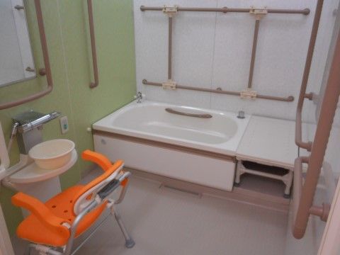 手前左にシャワー、カランとオレンジ色のシャワーチェア。奥に浴槽と大きなバスチェアが置かれている。壁の全面に手すりが張り巡らされている。