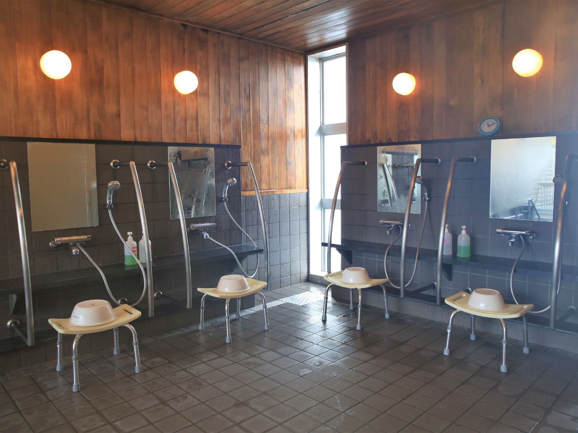 ダークグレーのタイルが壁と床に貼られた浴室で、壁の半分は板張りになっている。洗い場は４か所にあり、それぞれに椅子と洗面器がある。