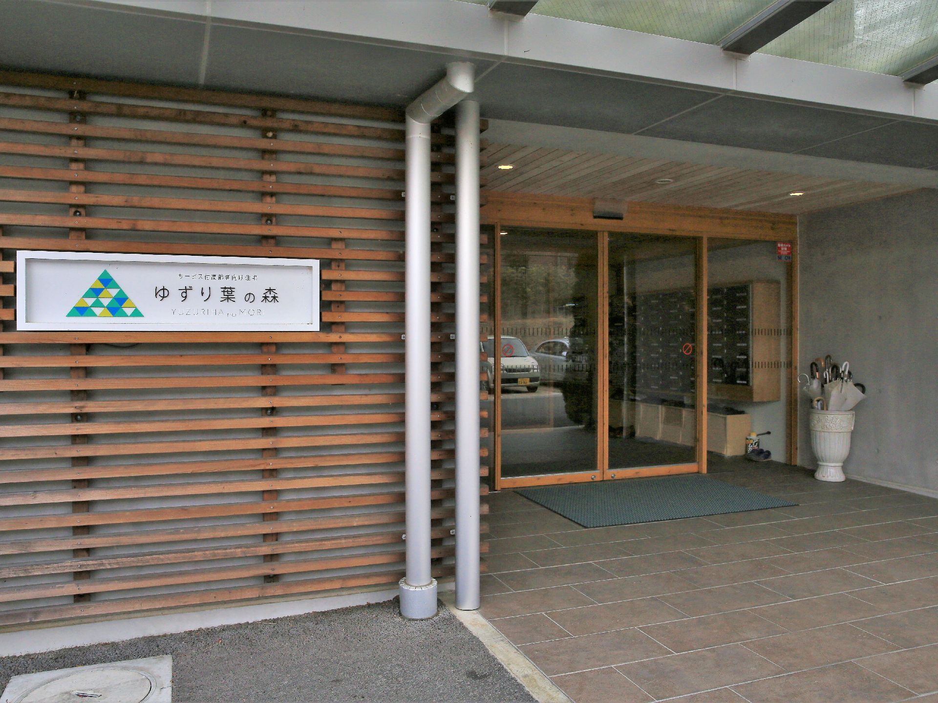 玄関扉は木材の枠がはめられたガラスの自動扉になっている。床にグレーの玄関マットが敷かれている。入口横に施設名が書かれた看板がある。
