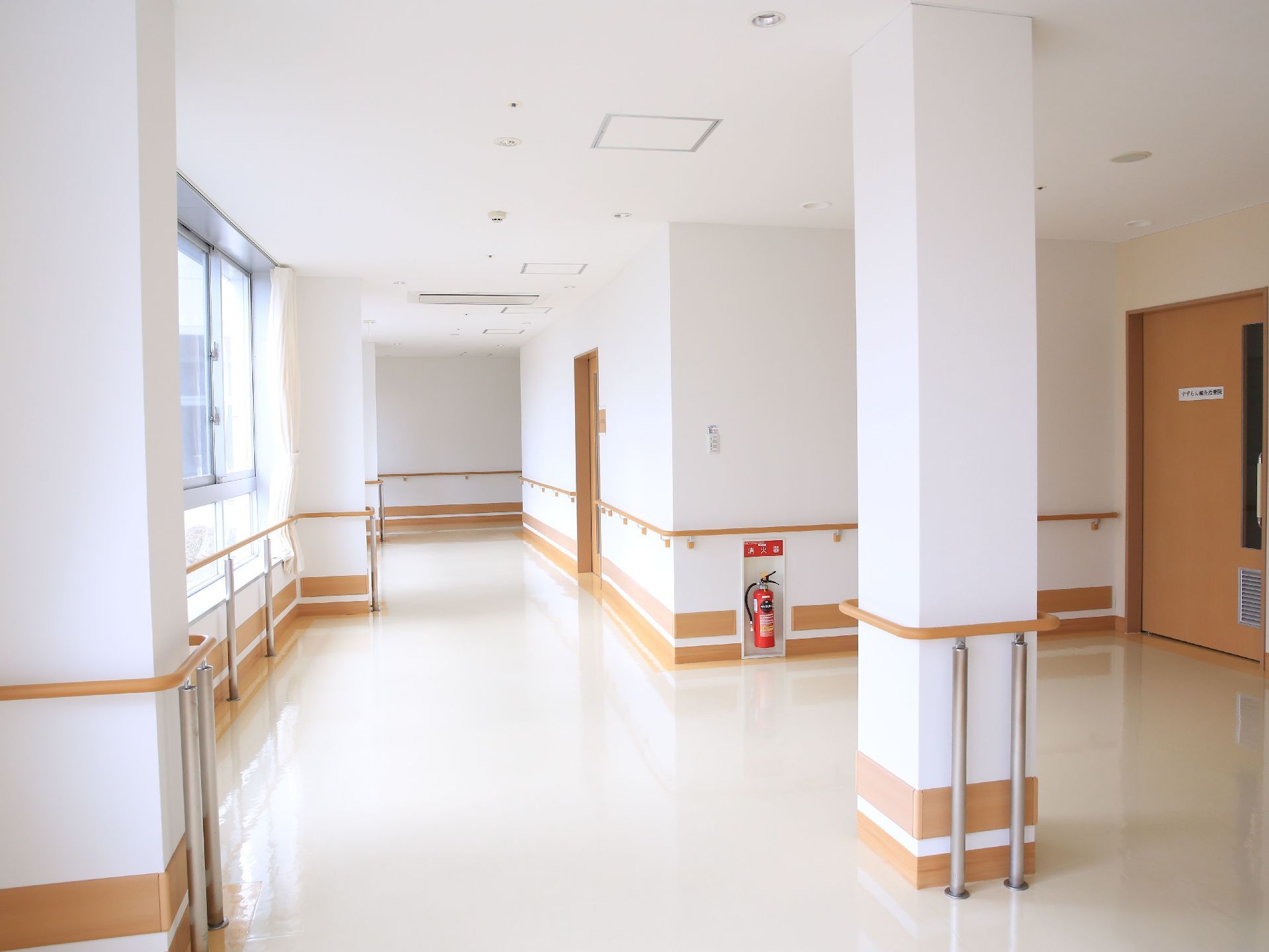 白い壁に木製の手すりが壁全面に取りつけられている。右手の奥に木製の扉が１枚ある。壁に消火器が設置されている。