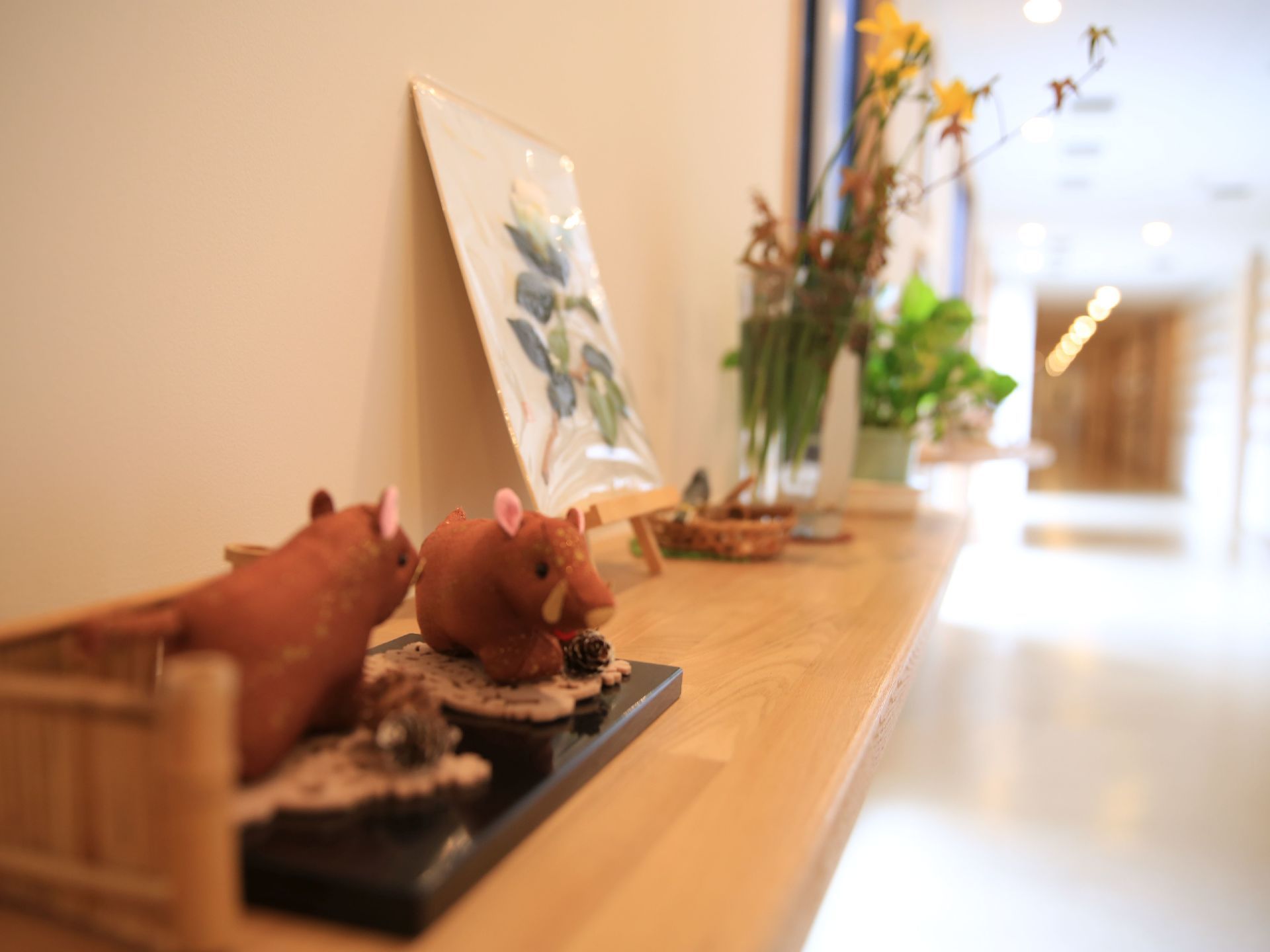 壁に作りつけられた棚に干支のオブジェや色紙の作品などが飾られている。奥に花瓶に生けられた花や鉢植えがある。