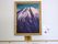 白い壁に、金色の額縁に入れられた大きな雪山の絵が飾られている。絵の下に作品名が貼られており手すりが取りつけられている。