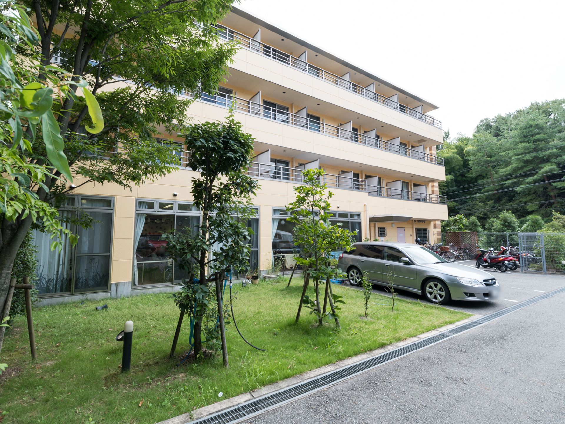 芝生の上に木が何本も植えられている。自動車が1台、自転車やバイクが複数駐車してある。建物は4階建てである。