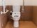 白色と深みのある木目仕様の内装のトイレで、横の壁際にはエル型の手すりとナースコールが設置してある。後ろの壁にも手すりがある。
