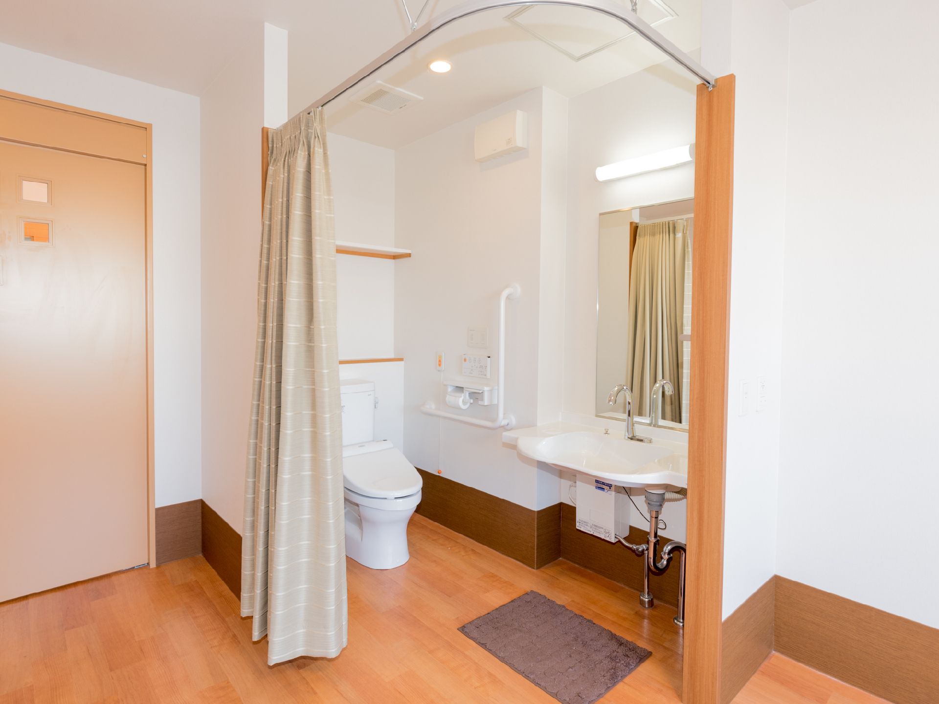 居室には、カーテンで仕切られたトイレがある。同じスペースに洗面台も設置されている。壁には、L字型の手すりや棚、鏡が取り付けられている。