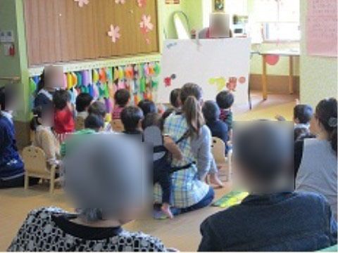 大型絵本の読み聞かせを聞いている園児と、後ろ姿の白髪の2人の人物