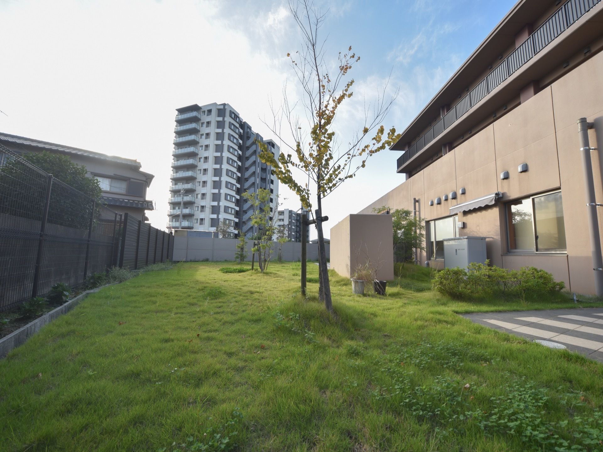 建物の隣に芝生敷かれ、木が植えられている。柵の前にも植物が植えられている。近隣はマンションなどの住居が建ち並んでいる。