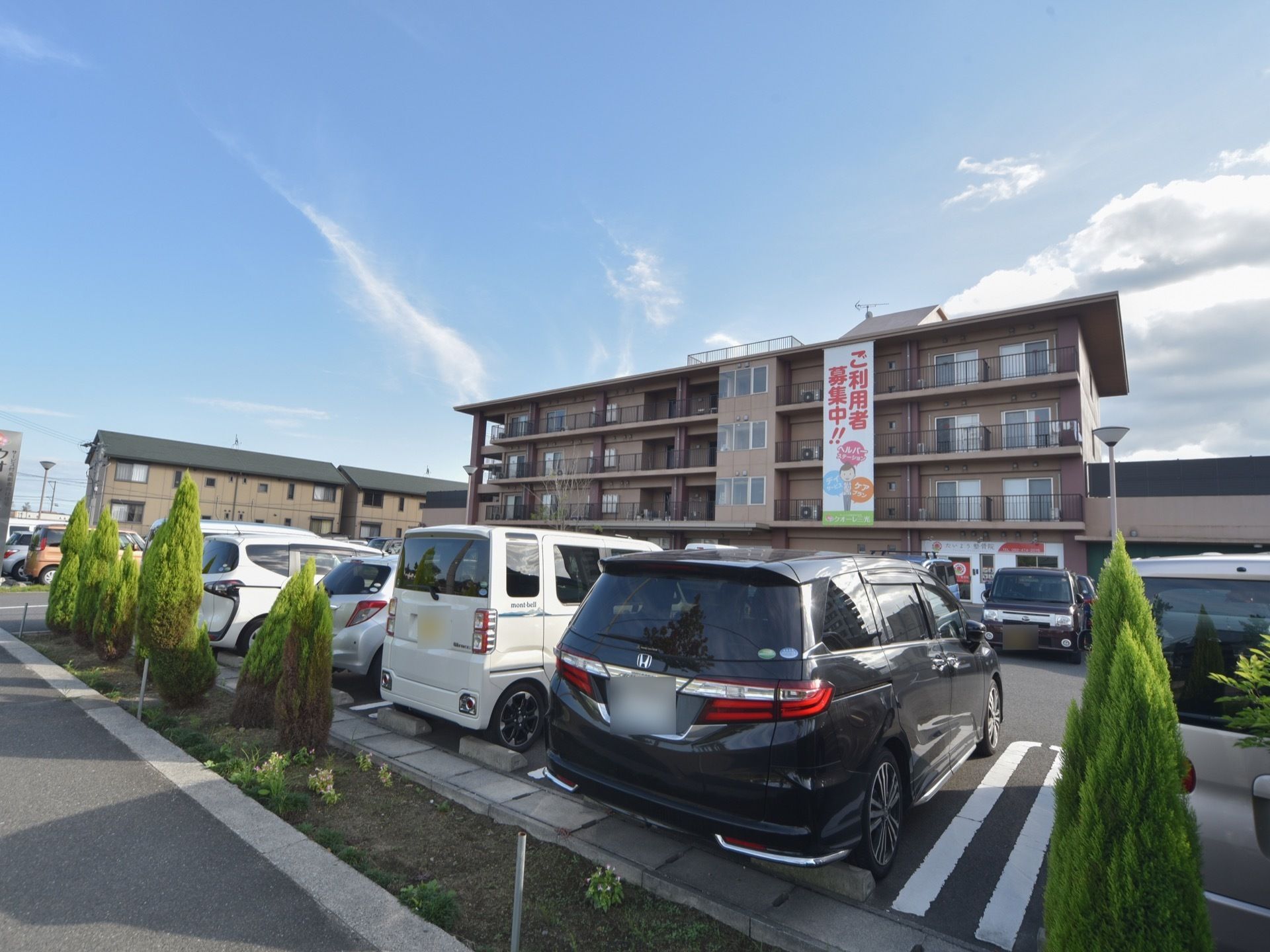 ４階建ての茶色の外壁の建物で、施設前に駐車場が完備されている。居室にはベランダがある。入居者様募集の表示がある。