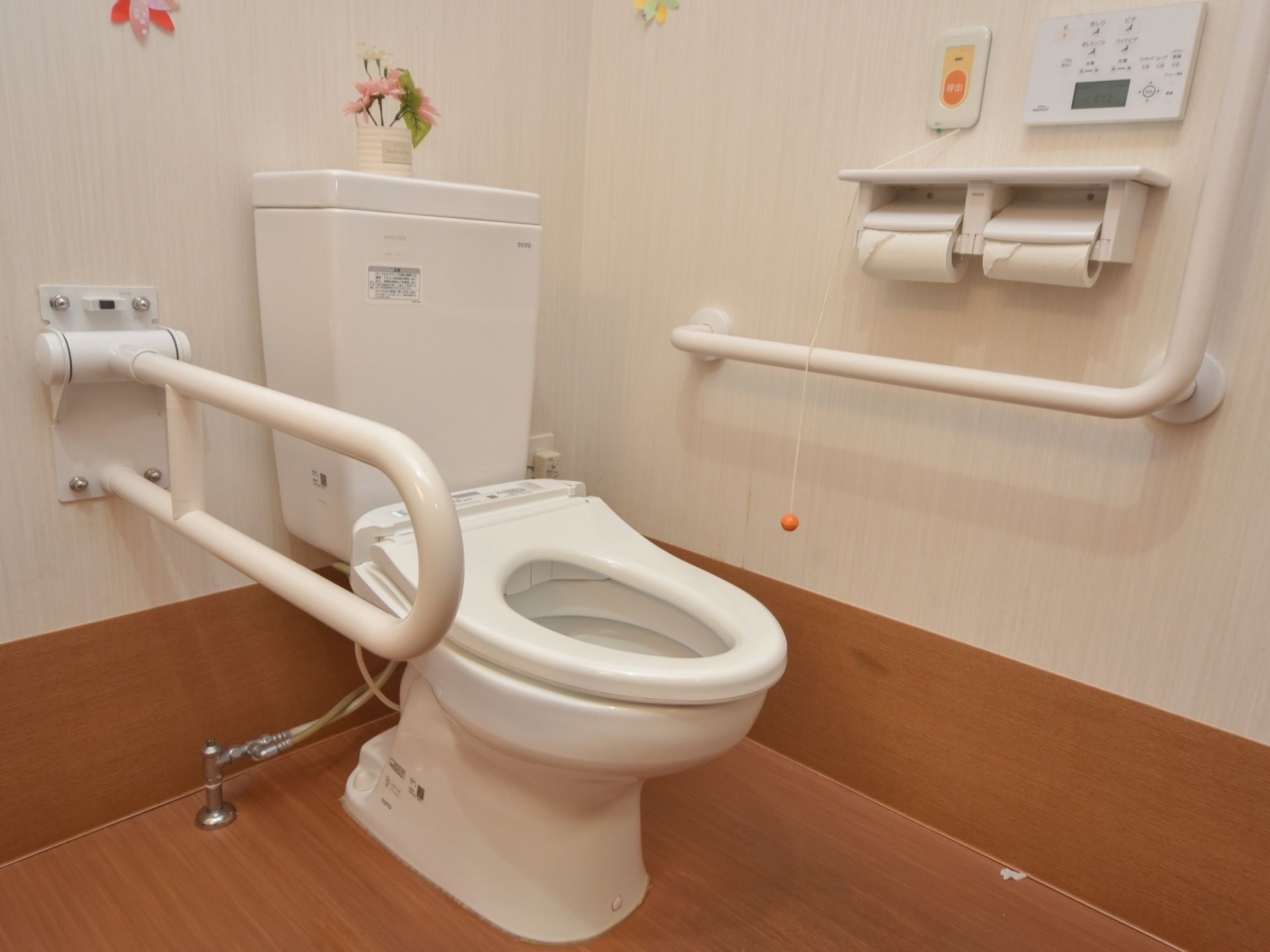 二種類の手すりが設置されたトイレである。花や壁の飾りがされている。オレンジ色のボタンと、そこからつながる紐は、緊急時呼び出しブザーである。