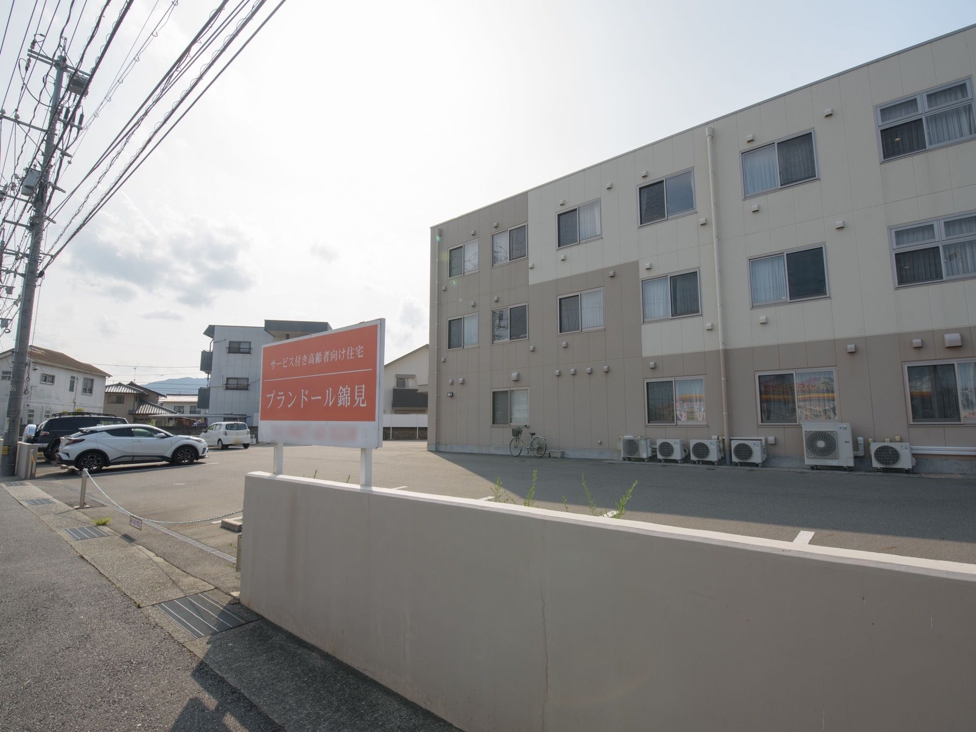 外壁が白色と灰色の３階建ての施設である。建物前には、アスファルトで整地された専用の駐車スペースがある。