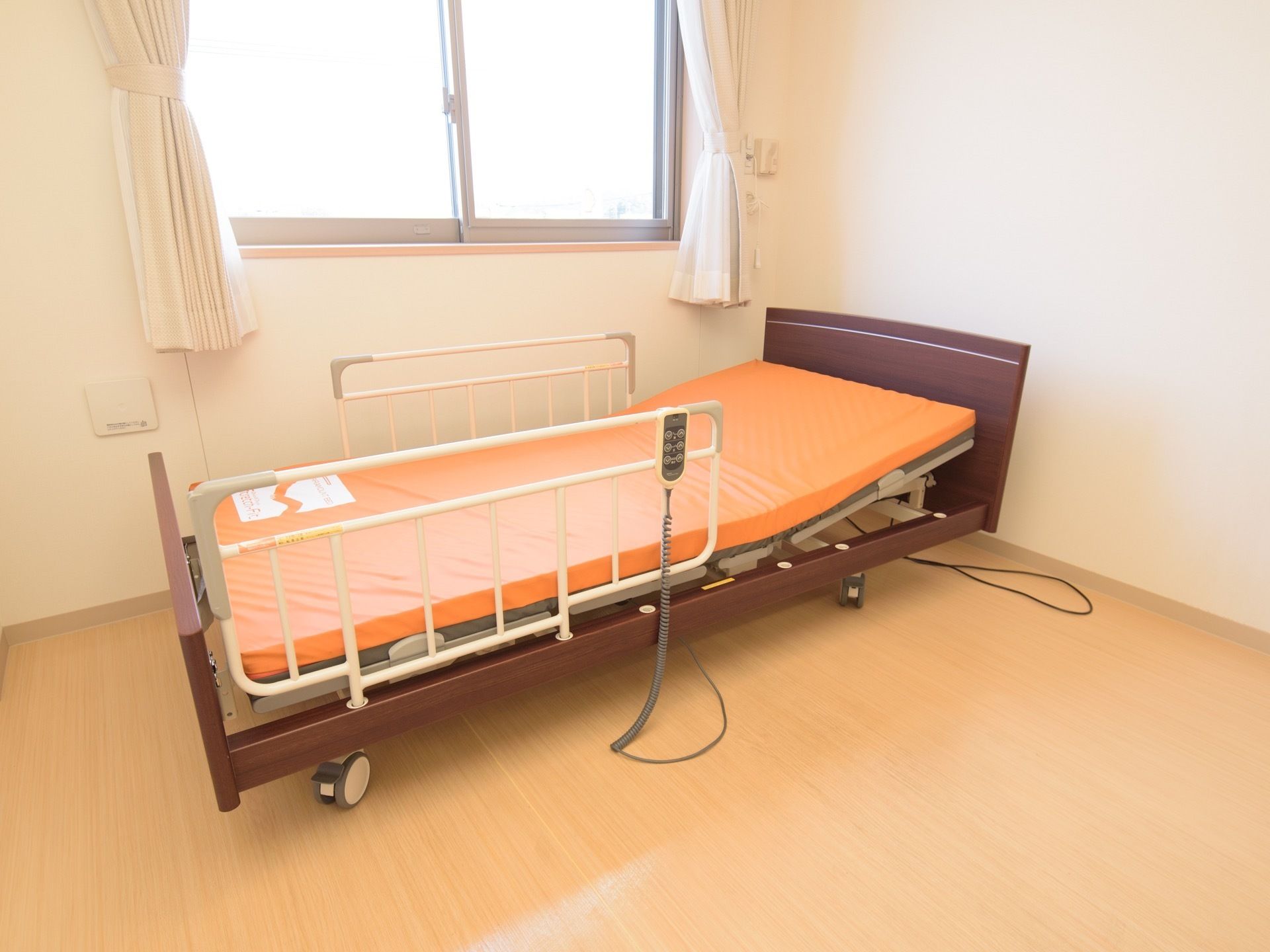 白色のカーテンが吊るされた腰高窓のある居室で、電動ベッドが置かれている。床は木目調のフローリングである。