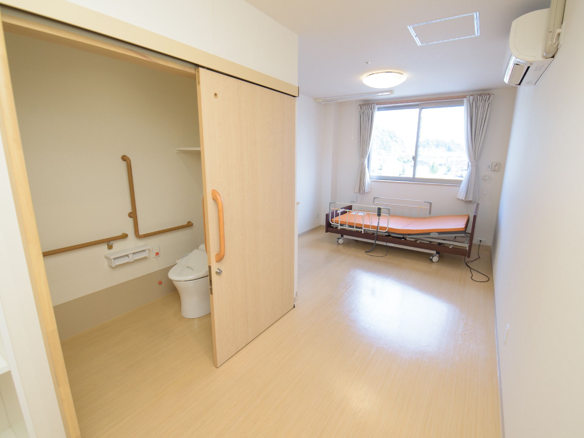 左の開けられたドアの向こうにトイレが見え、奥の窓際にベッドが置かれた居室