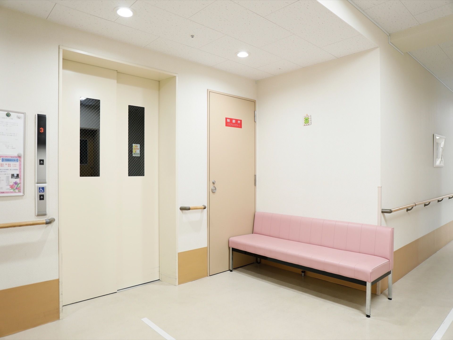 施設内に白いエレベーターがある。右横には非常時の扉やピンク色のベンチが置かれている。壁には手すりが付いている。