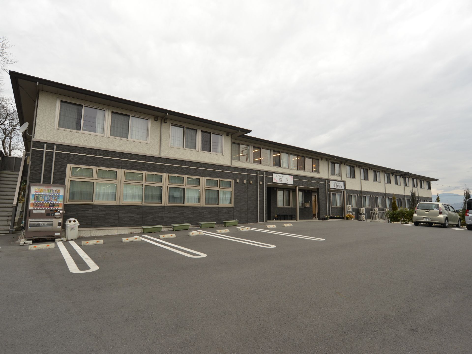 軽量鉄骨2階建ての建物。1階部分はグレー、2階部分は白のツートンカラーとなっている。建物の前には駐車スペースが十分ある。