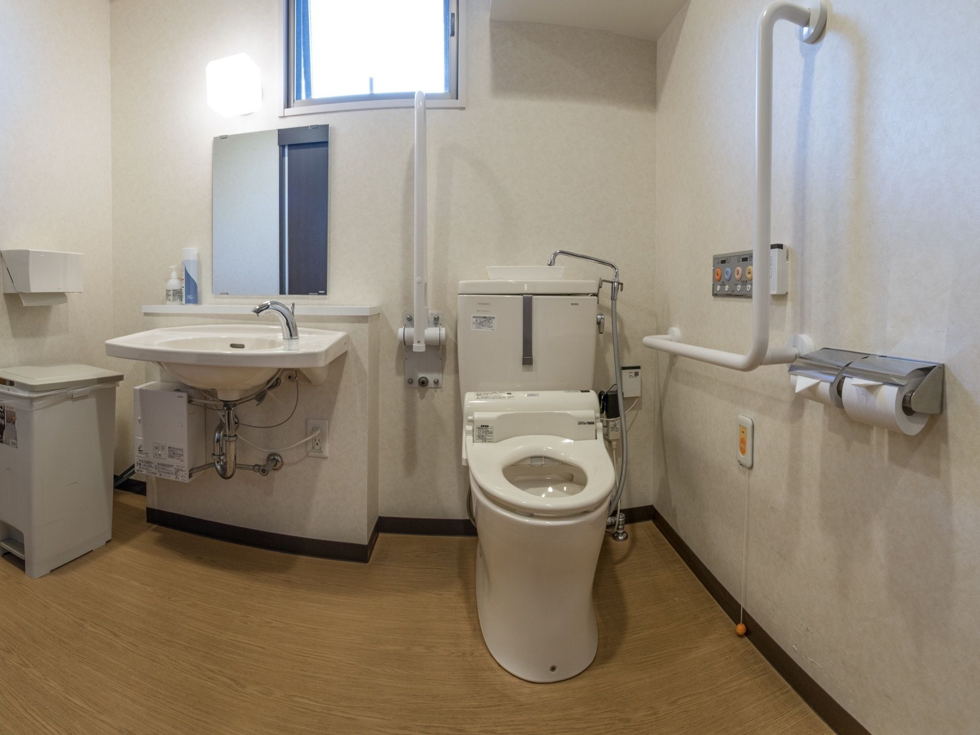 広いトイレには、タンク付きのトイレの隣りに洗面台とゴミ箱が設置されている。手すりもたくさん取りつけられている。