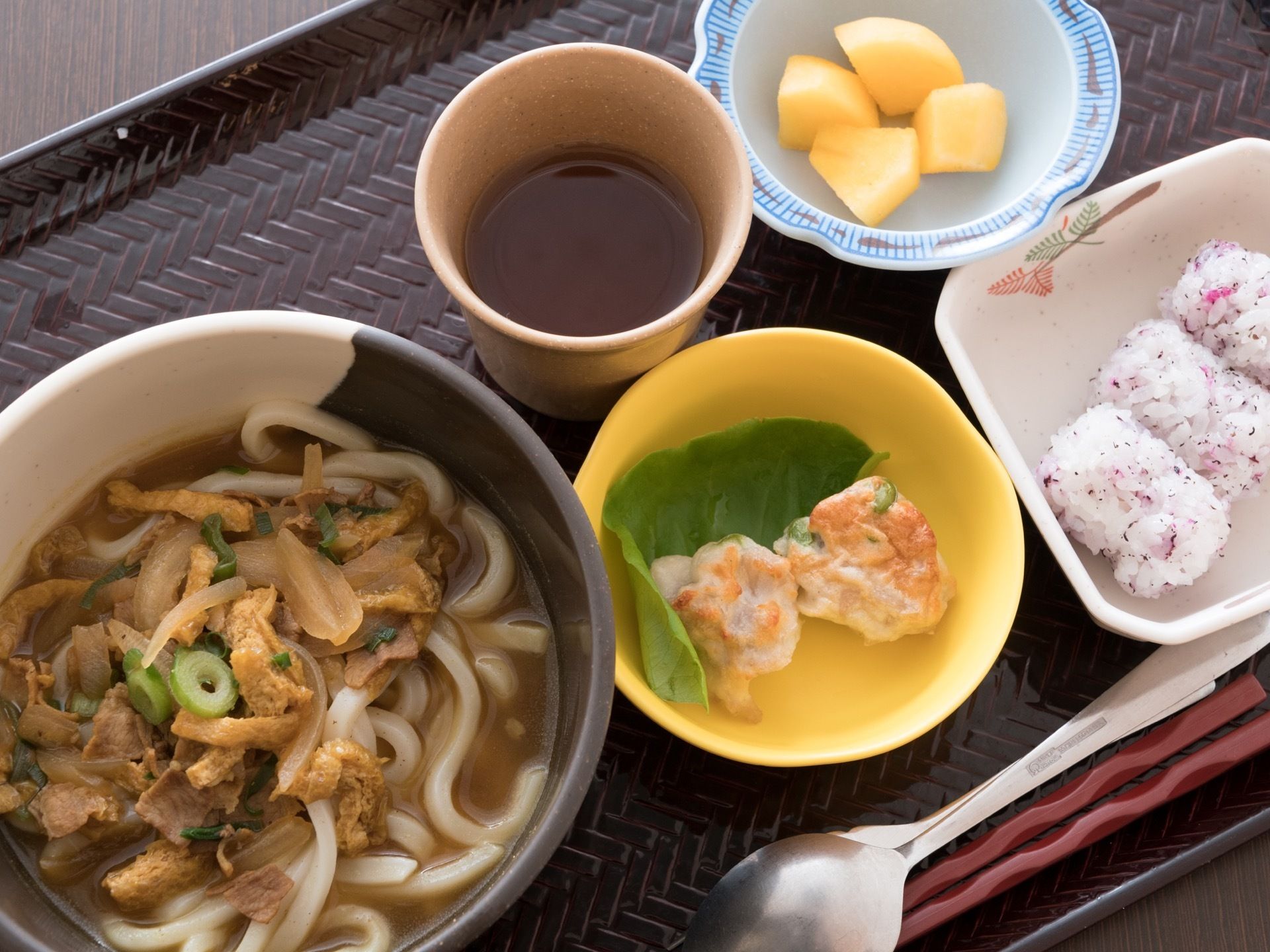 黒いトレーの上には、うどんやおにぎり、りんごなどの4種類の食事が器に盛りつけられている。お茶も添えられている。