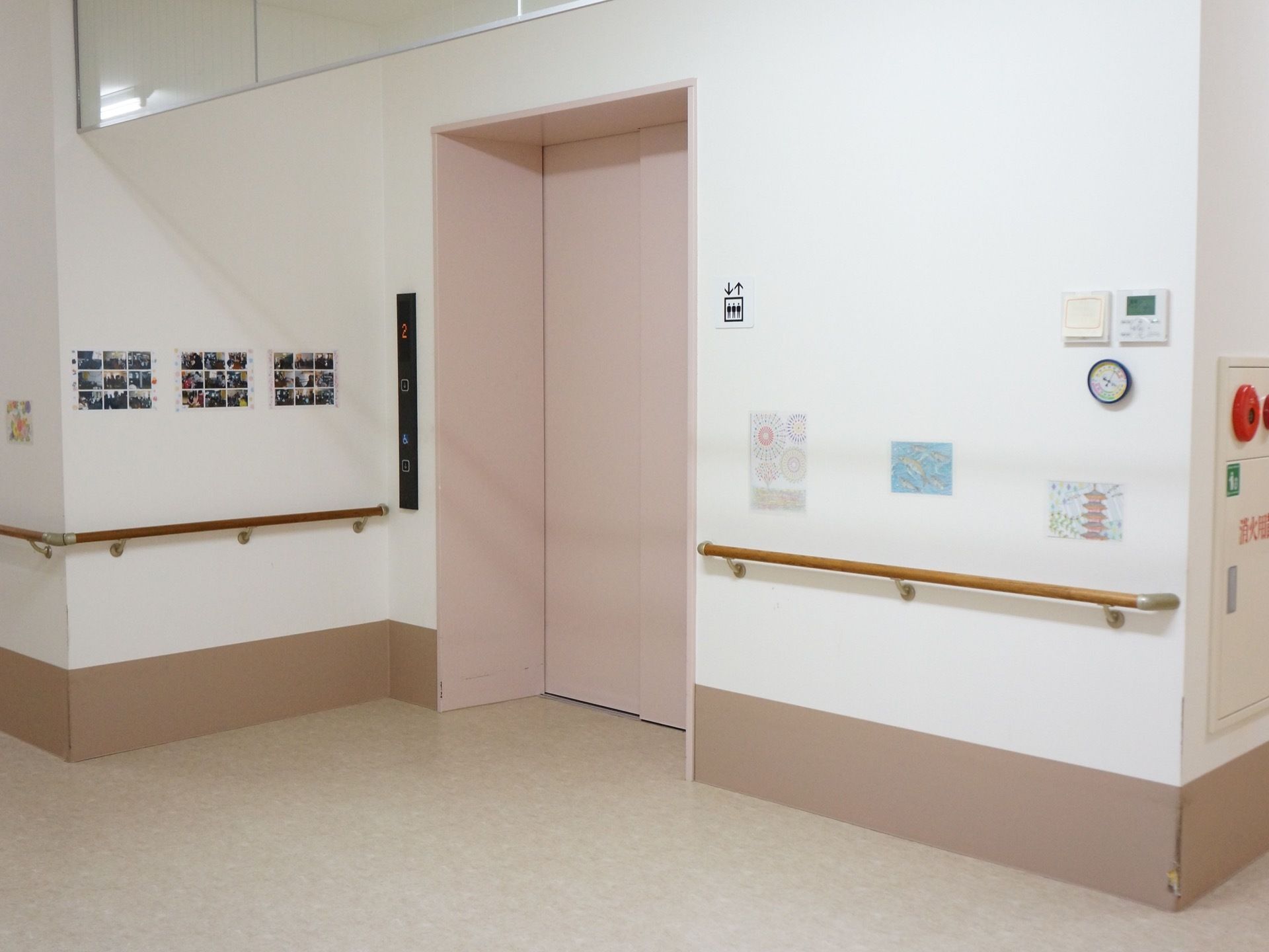 ピンクの扉のエレベーターが設置されており、エレベーター脇の白い壁には手すりが設置され、写真が貼られている。