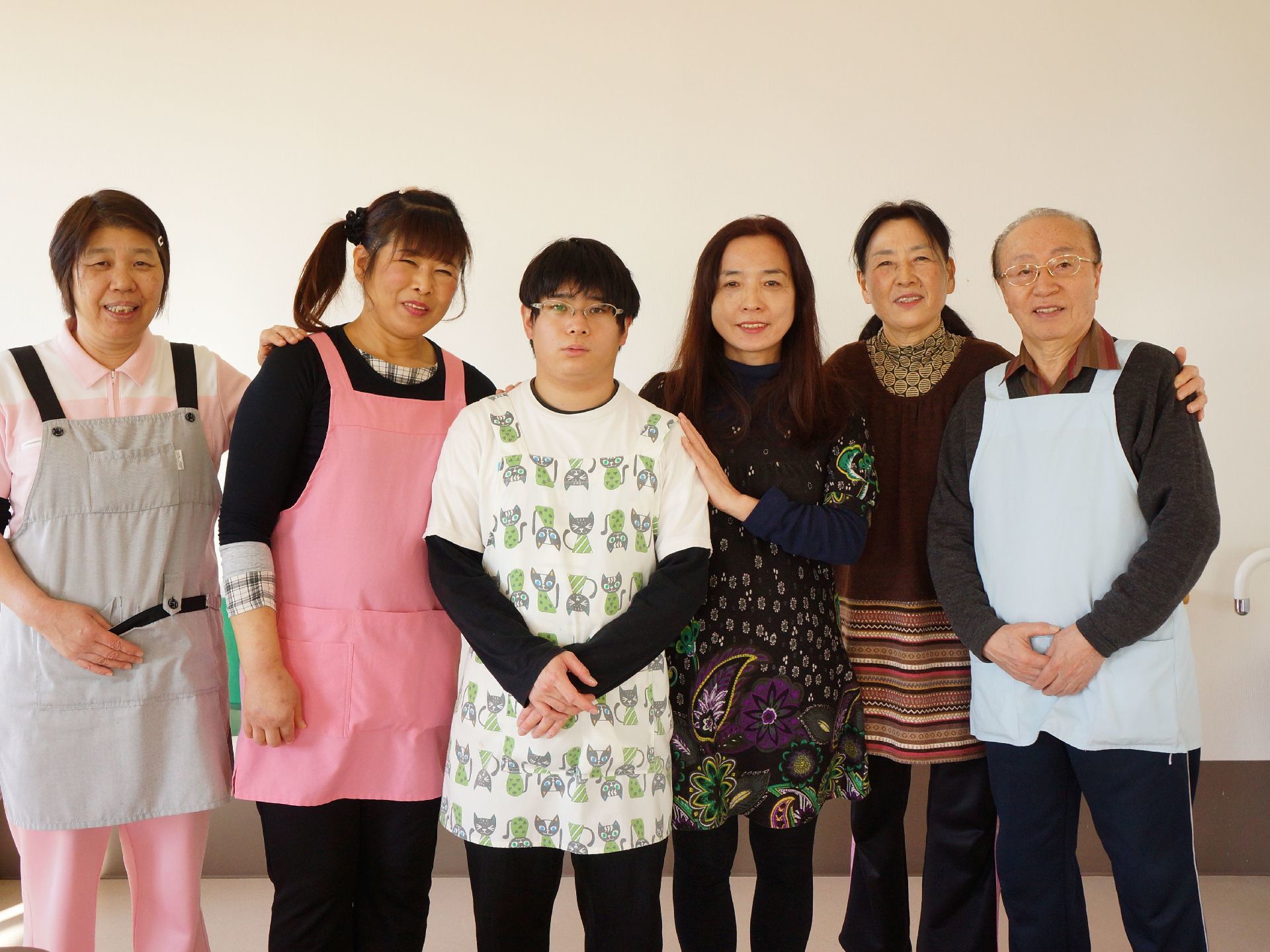 6人の男女が肩を寄せ合って並んで立っている。年齢はバラバラだが、にこやかな表情で和気あいあいとしている。