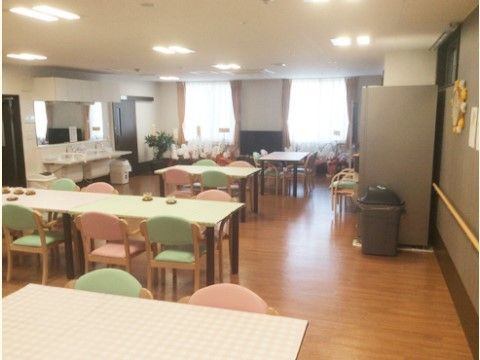 奥に窓、左奥に手洗いスペースのある、テーブルと緑とピンクの椅子が並んだ食堂