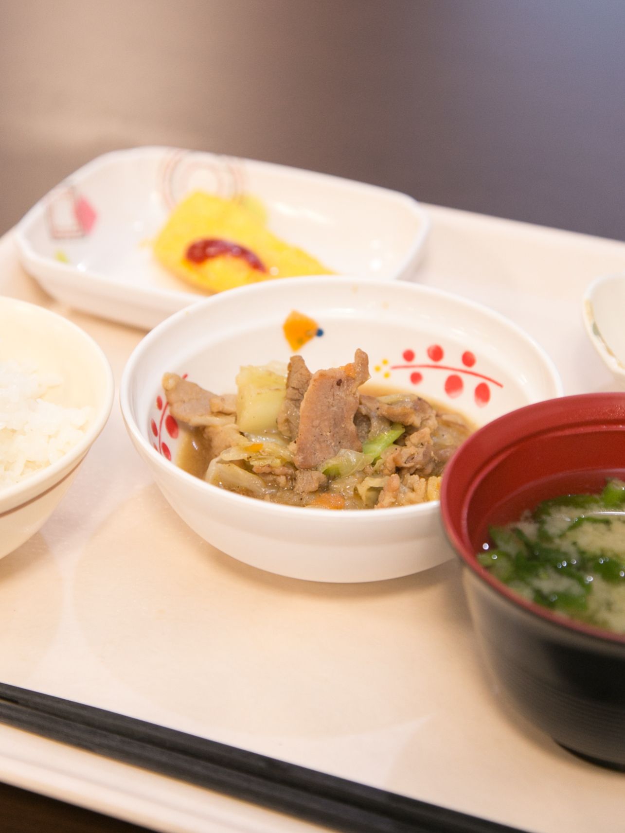 白い四角いトレーの上に食事がのっている。手前に箸とご飯、味噌汁が置かれていて、他におかずが３皿彩よく並べられている。
