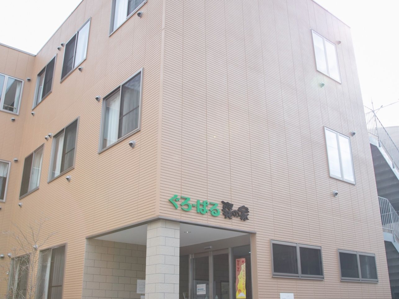 出入り口に階段とスロープが設けられていて、スロープには黄色い点字ブロックが敷かれている。建物の横に木が植えられている。
