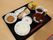 お吸い物やご飯、煮物、焼き鮭といった食事が黒いトレイの上に並べられている。箸やマグカップに入った飲み物も用意されている。