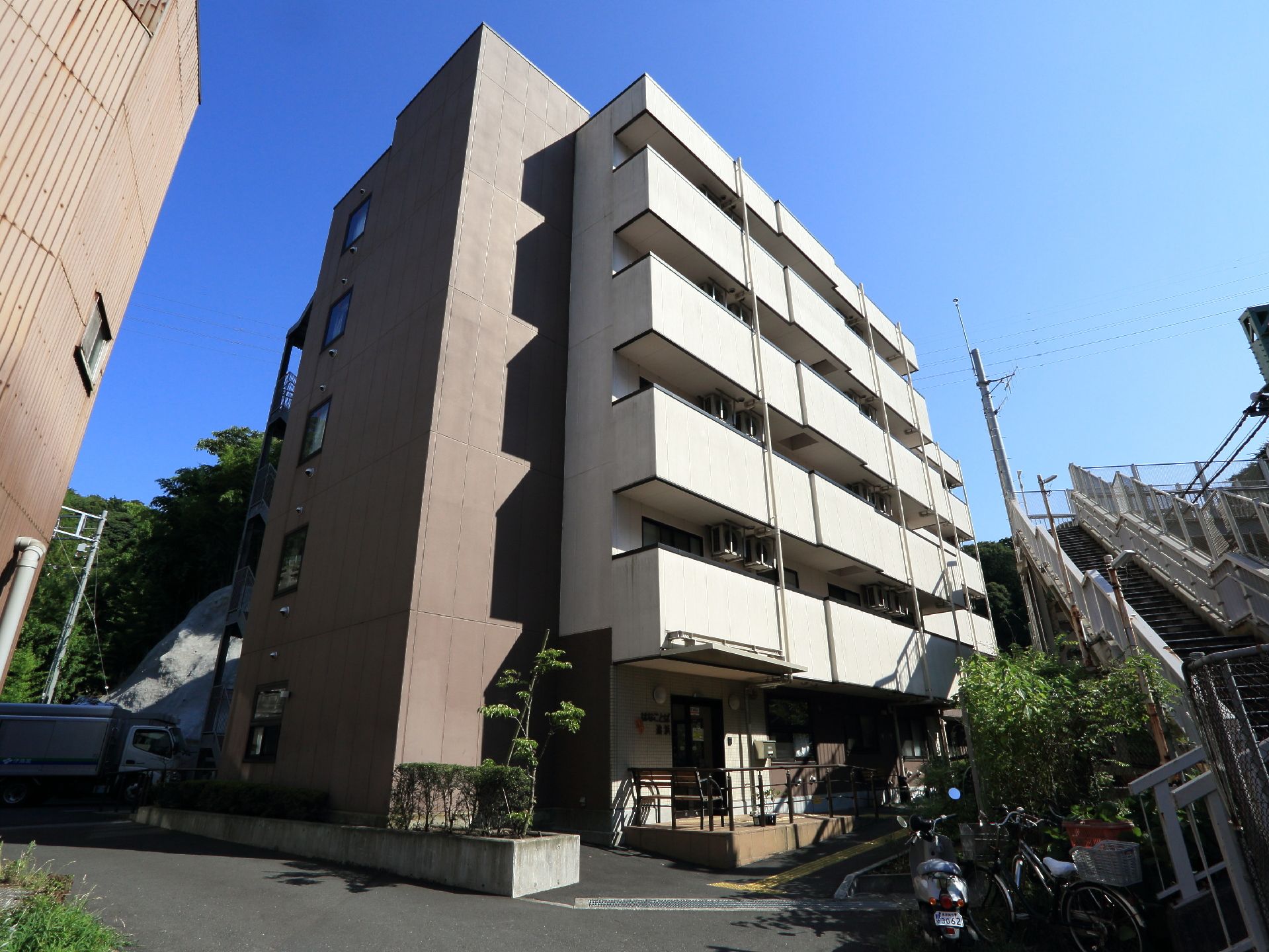 自然豊かな閑静な住宅街に建つ茶系と白色の外壁の鉄骨造の5階建ての建物で、建物横の所に植栽が施されている。