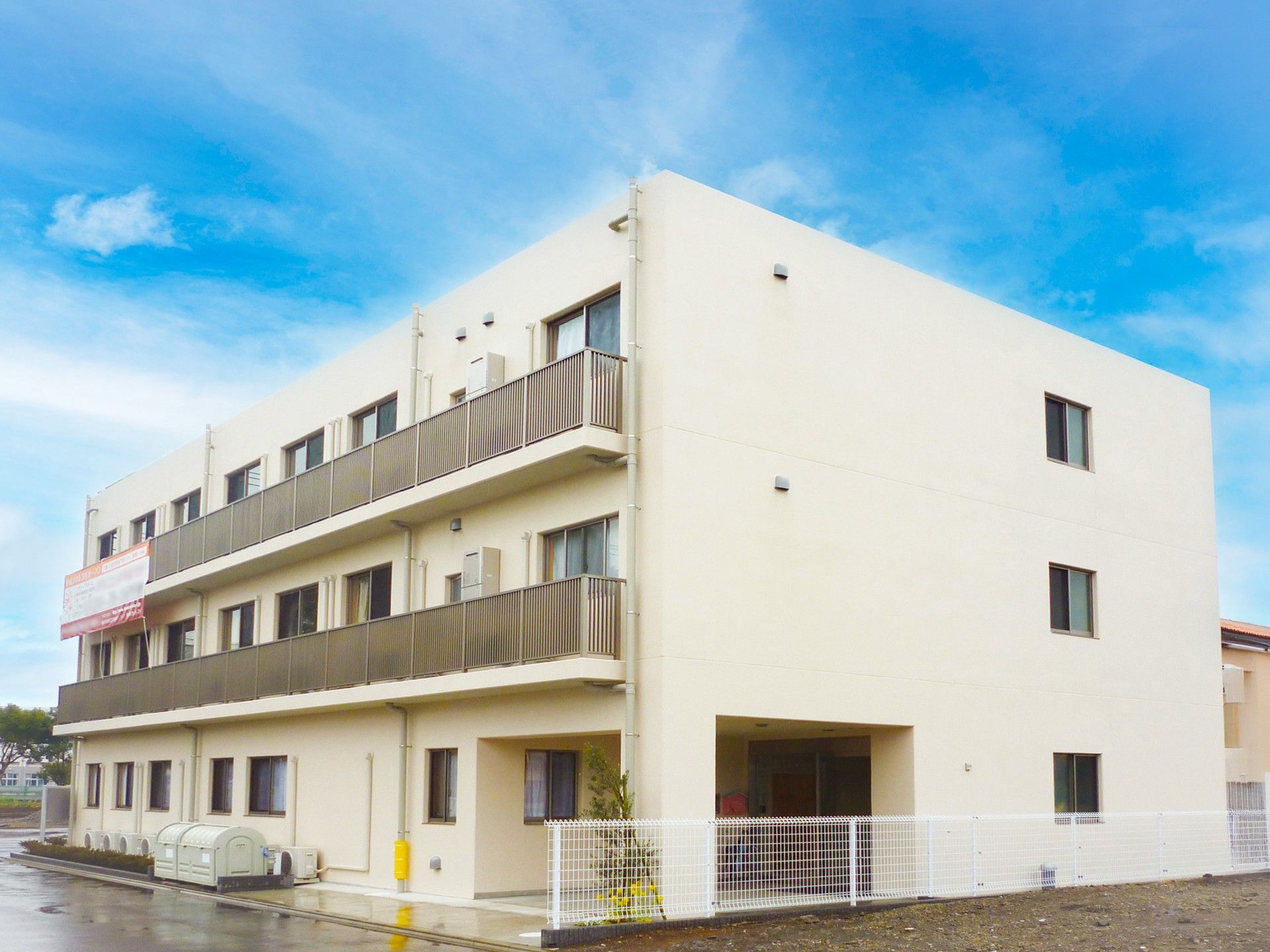 白を基調としたマンションタイプの外観である。住宅街の一角に位置しており、周りには花壇が設置されている。