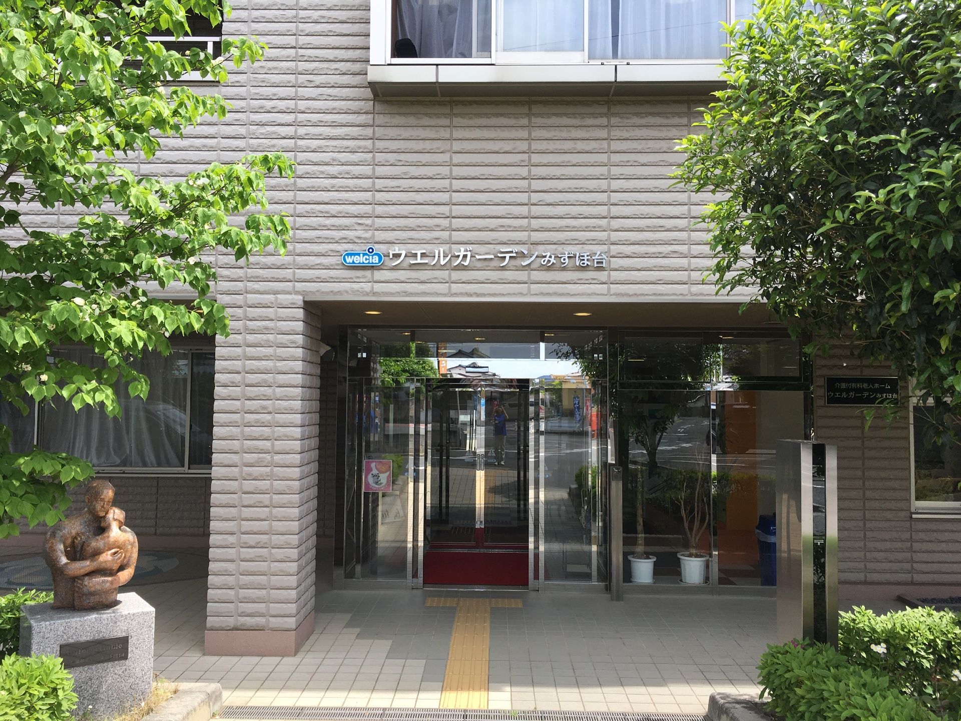 施設入口部分には青文字の入った銀色の看板が立てかけられている。建物付近には大きい緑の木々が生い茂っている。