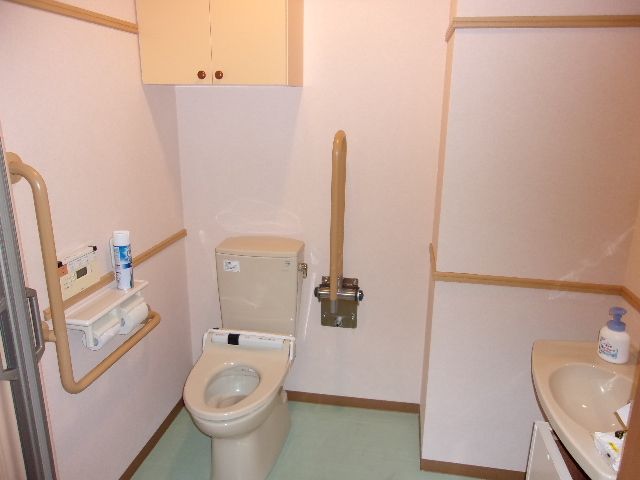 タンク付きの洋式トイレに可動式の手すりが設置されている。便器の頭上に収納棚と換気扇が取り付けられている。