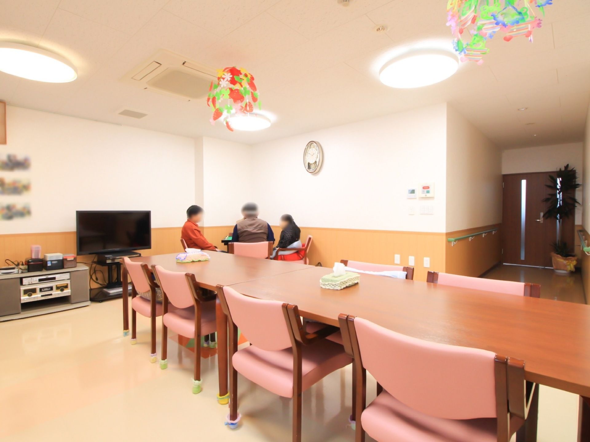 横長の木製テーブルが並べられた、共有スペースである。壁に腰板が貼られている。テレビやオーディオ機器もある。