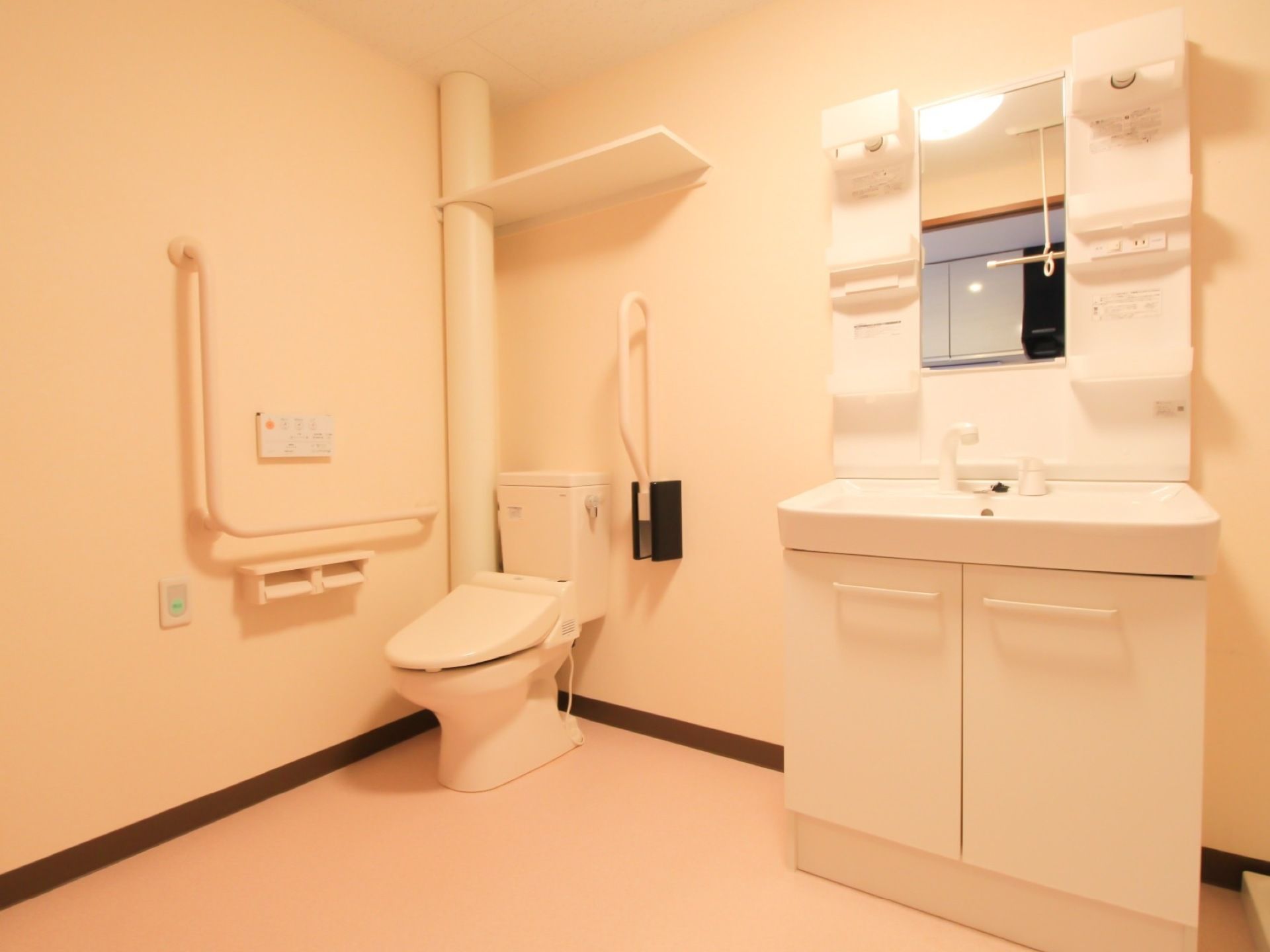 あたたかい色合いのトイレである。洋式便座が完備されている。Ｌ型の手すりがあるほか、上部には収納棚が設置されている。