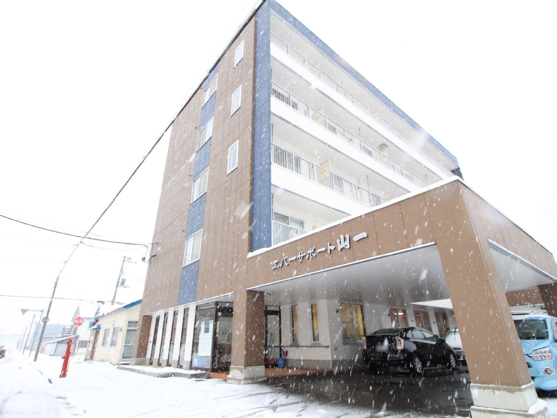 マンション風の施設外観である。5階建ての建物であり、落ち着いたブラウンで施されている。車が何台が停められている。