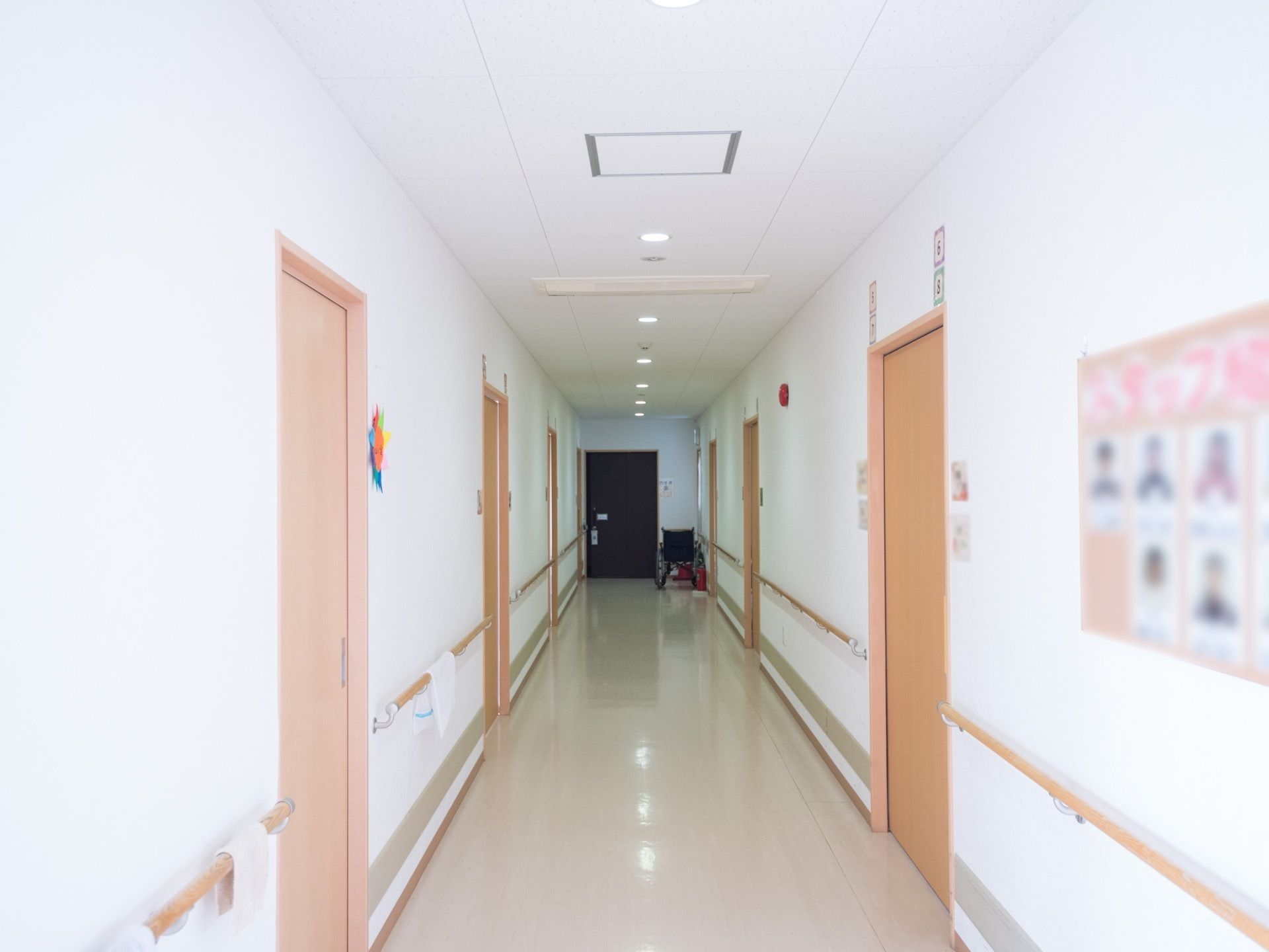 白を基調とした通路の奥には黒いドアがあり、側には車椅子が置かれている。壁の両側には居室のドアが並んでいて、手すりが設置されている。