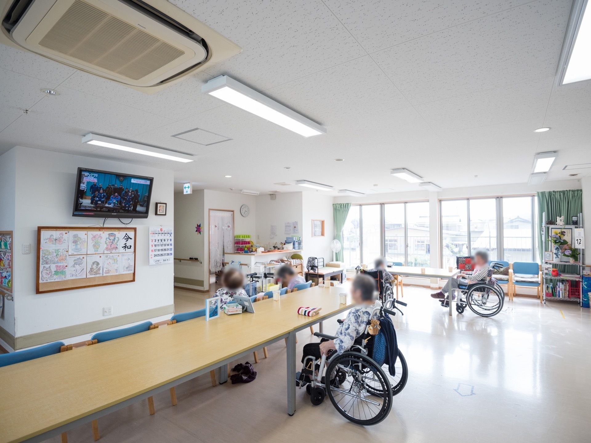 広々として明るい共有スペースには、長いテーブルや椅子が置かれている。壁にはテレビやカレンダーなどが掛けられている。