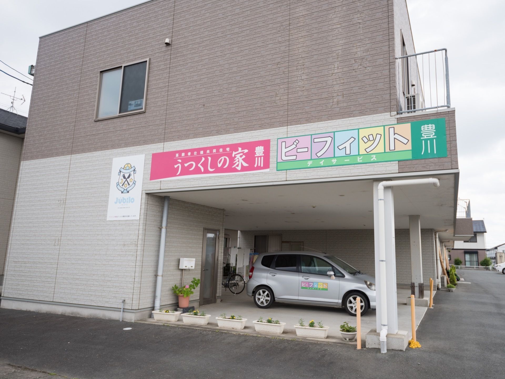 2階建ての1階部分には駐車スペースが設けられ、施設の車が停められている。壁には施設とデイサービス2つの看板が付いている。