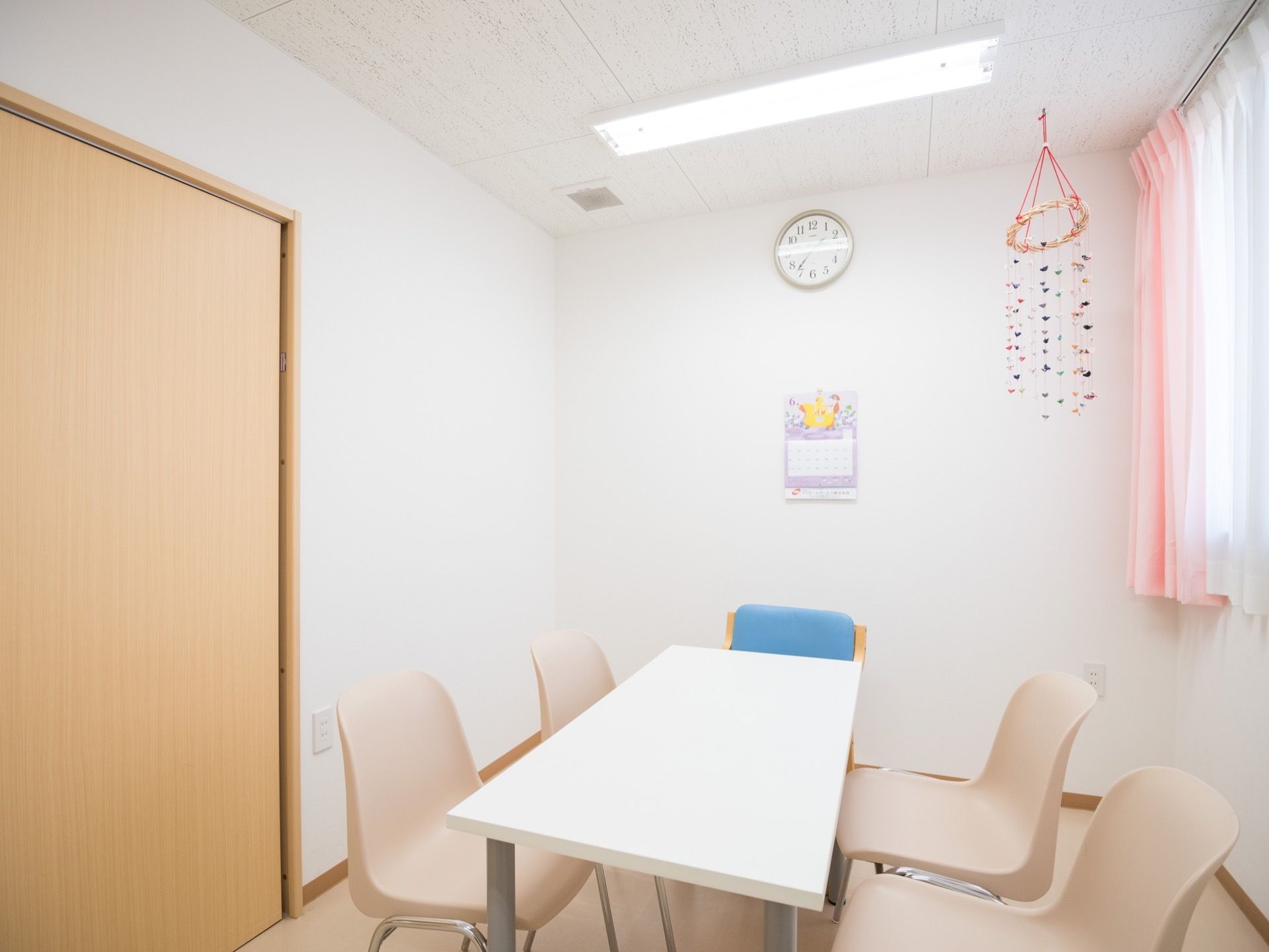 白い壁の個室で、天井近くに時計とカレンダーが掛けられている。中央にテーブルがあり、椅子が５人分置かれている。