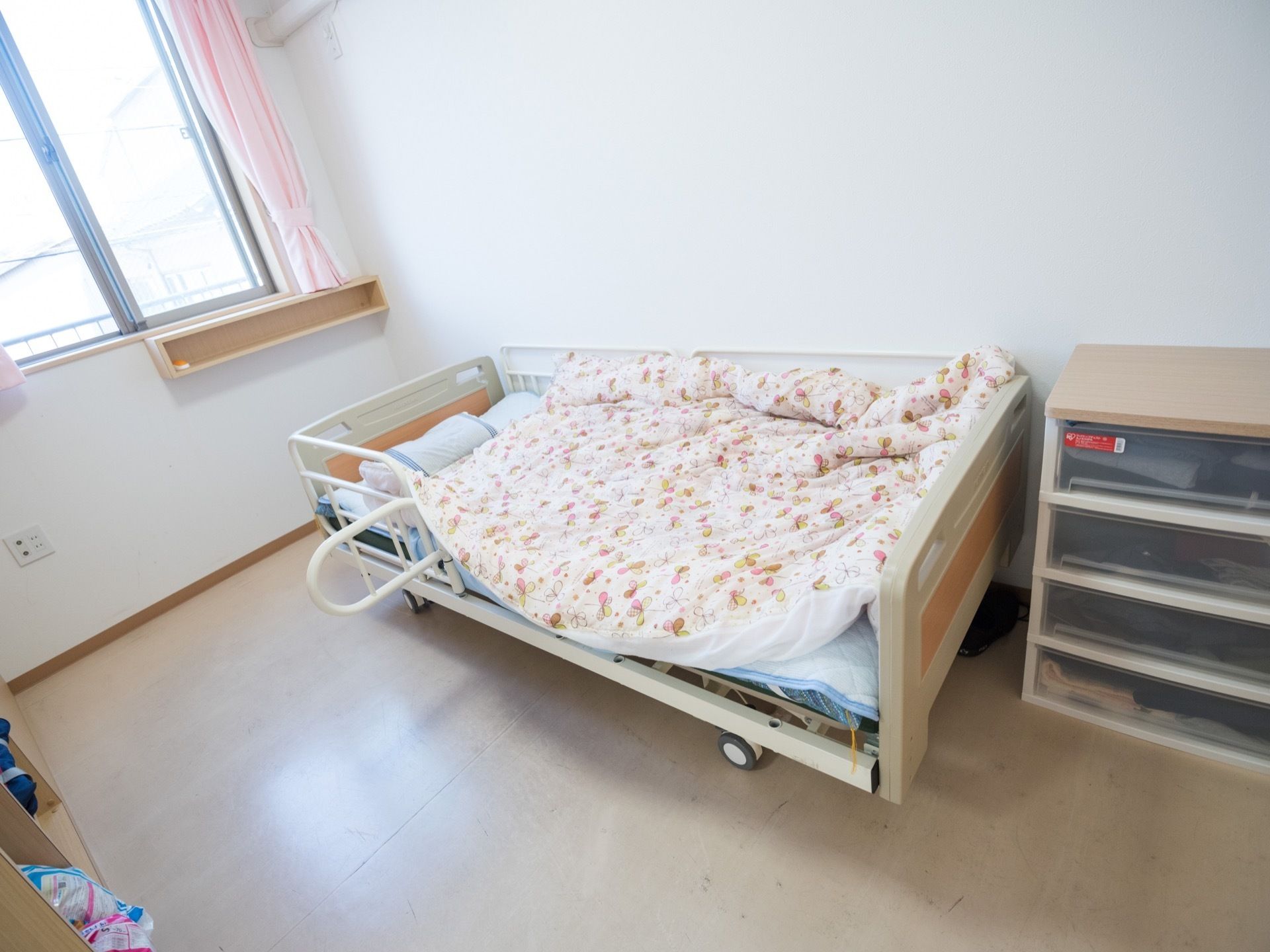 居室には大きな窓があり、ピンク色のカーテンが掛けられている。壁際に介護用ベッドが置かれ、花柄の布団が敷かれている。