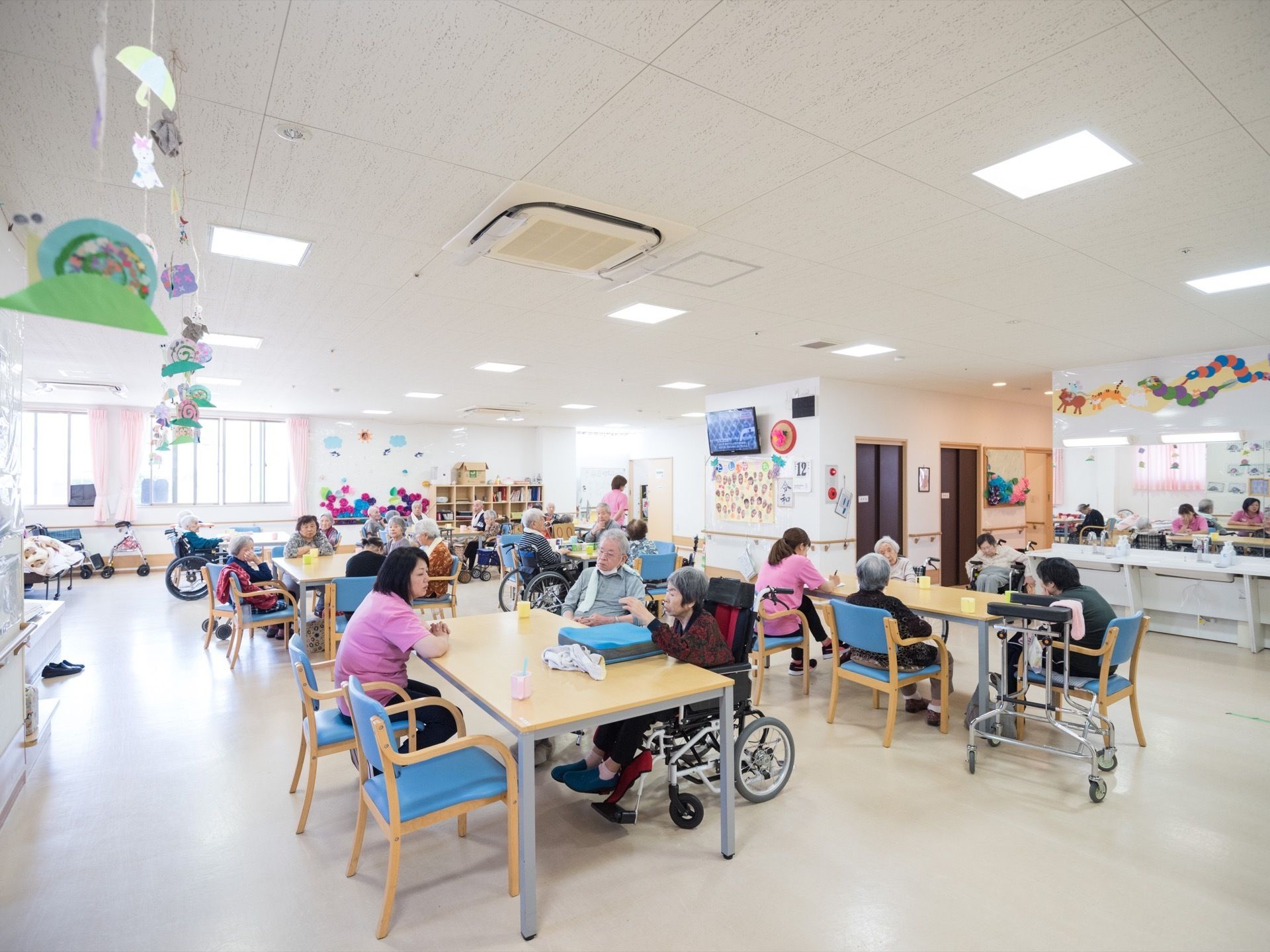 広々とした広間にたくさんのテーブルセットが並べられている。スタッフと入居者様が座っている。壁に液晶テレビが設置されている。
