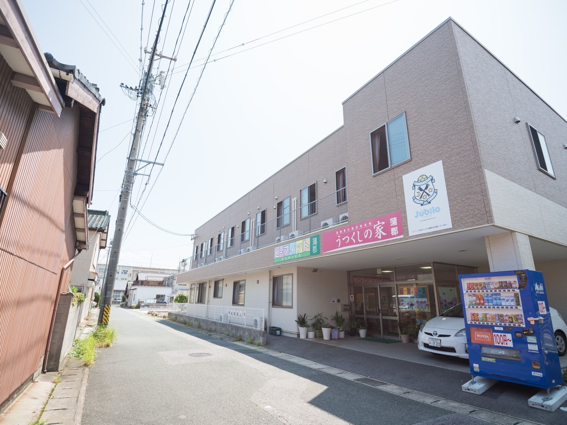 薄い紫色の２階建ての建物で、施設前は道路が通っている。１階部分は駐車スペースと玄関があり、自動販売機が設置されている。