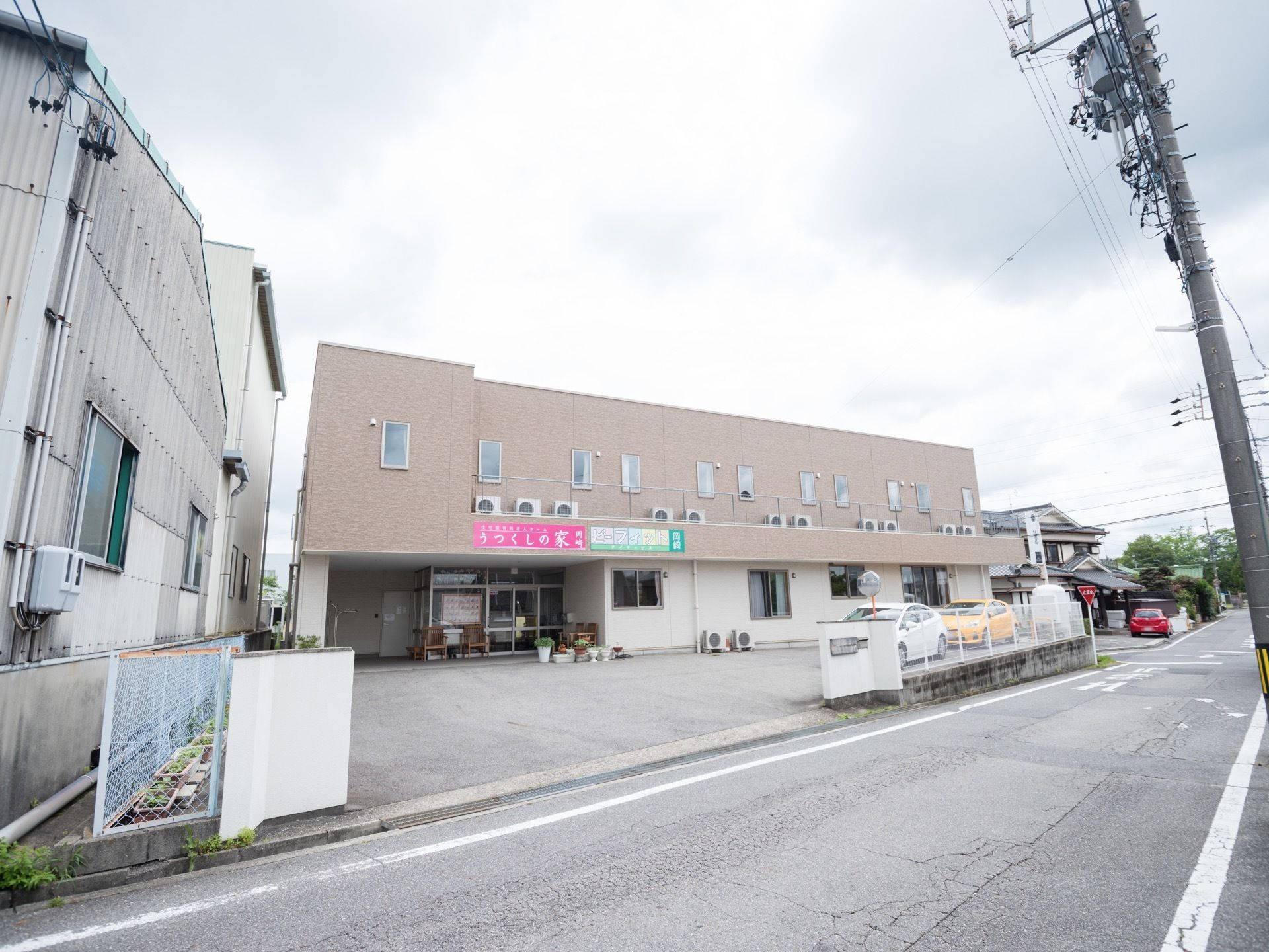 住宅街の中に２階建ての建物が建っている。ベージュを基調とした柔らかな雰囲気の建物で、敷地内に車が３台駐車している。