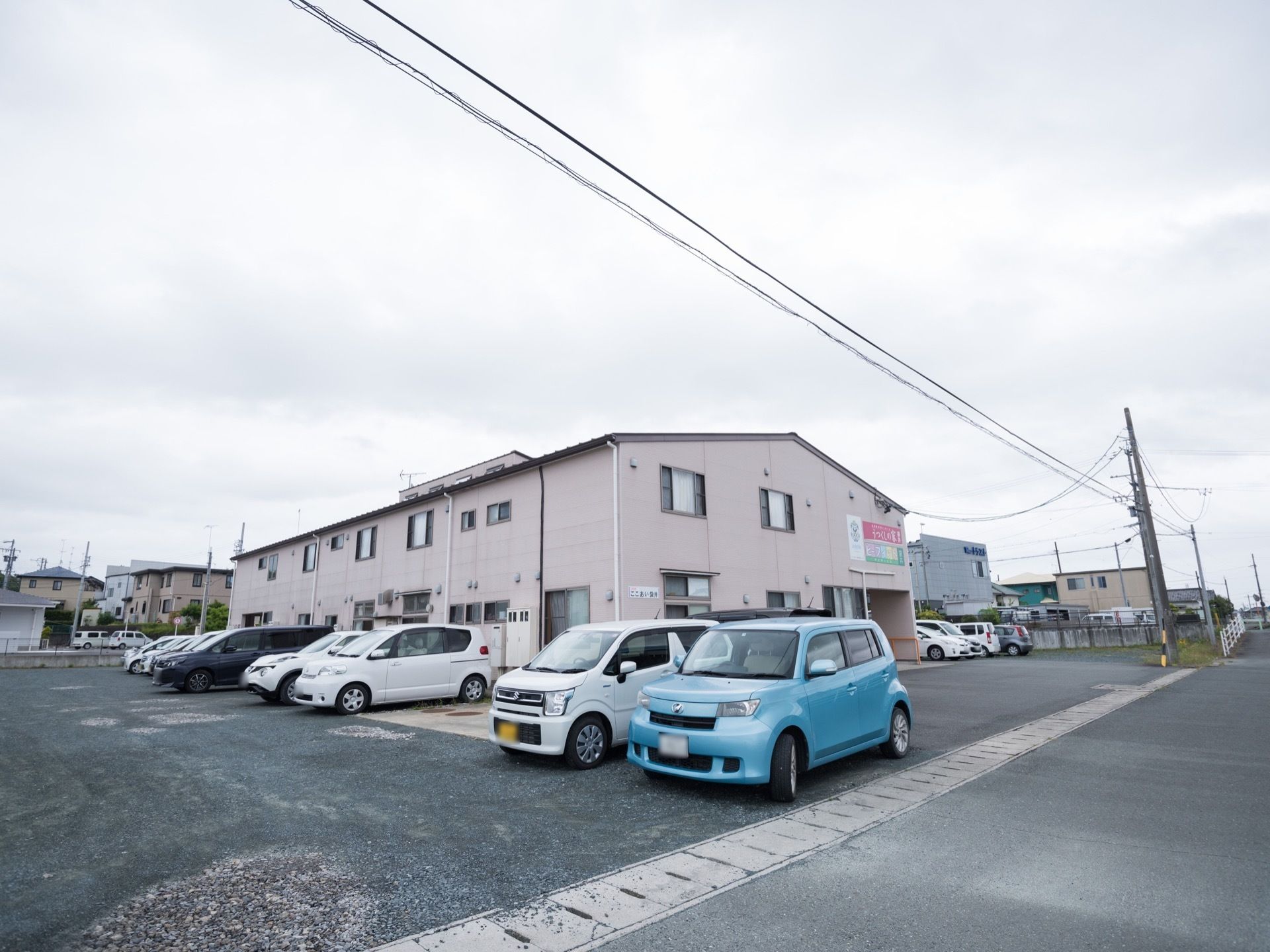 広い道路に面した敷地に2階建ての建物が建っている。横は舗装された駐車場になっていて、車が十数台停められている。