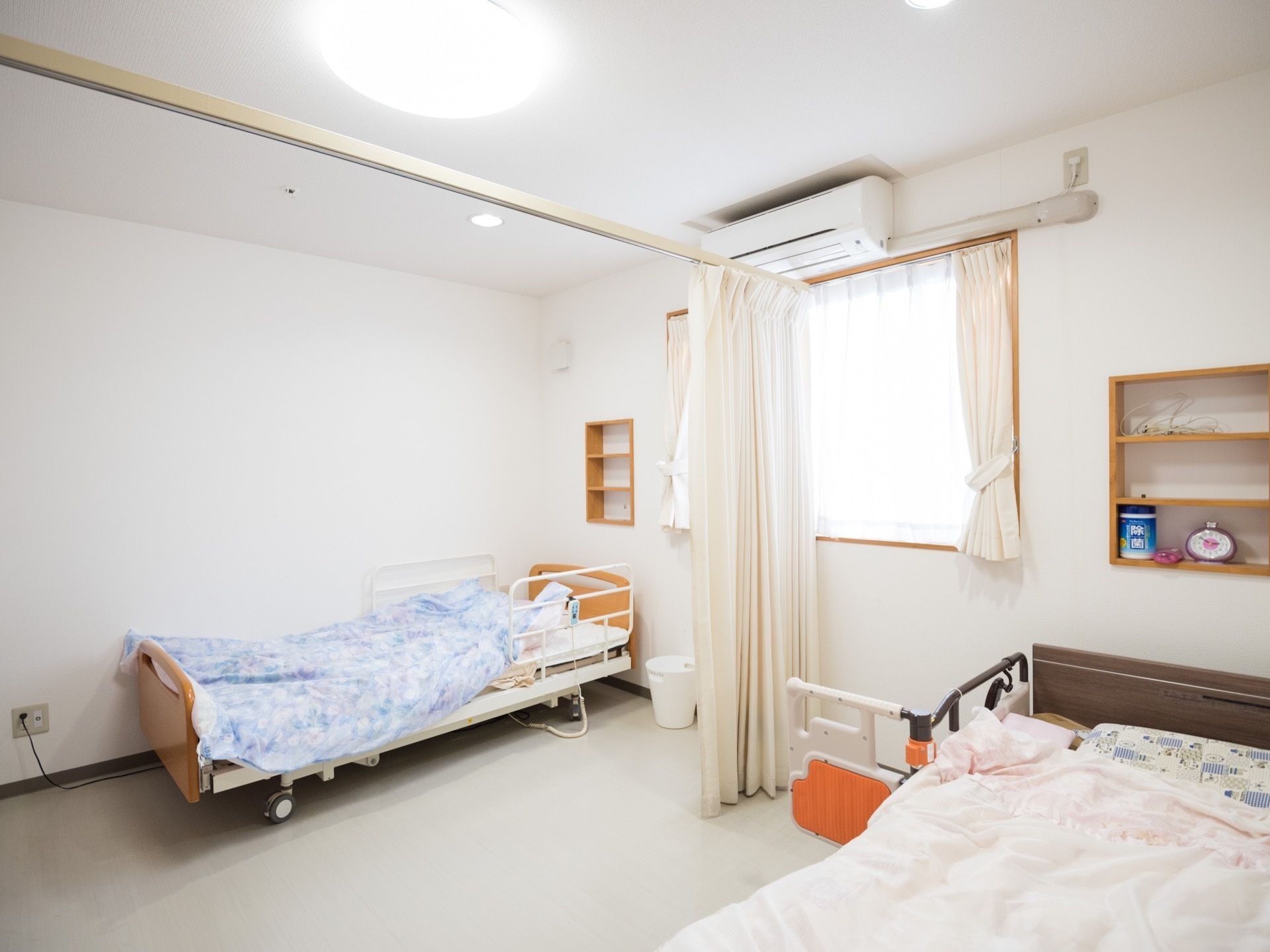 居室内にベッドが2台設置されている。中央を仕切るようにカーテンレールが敷かれている。窓があり、その上にはエアコンがある。