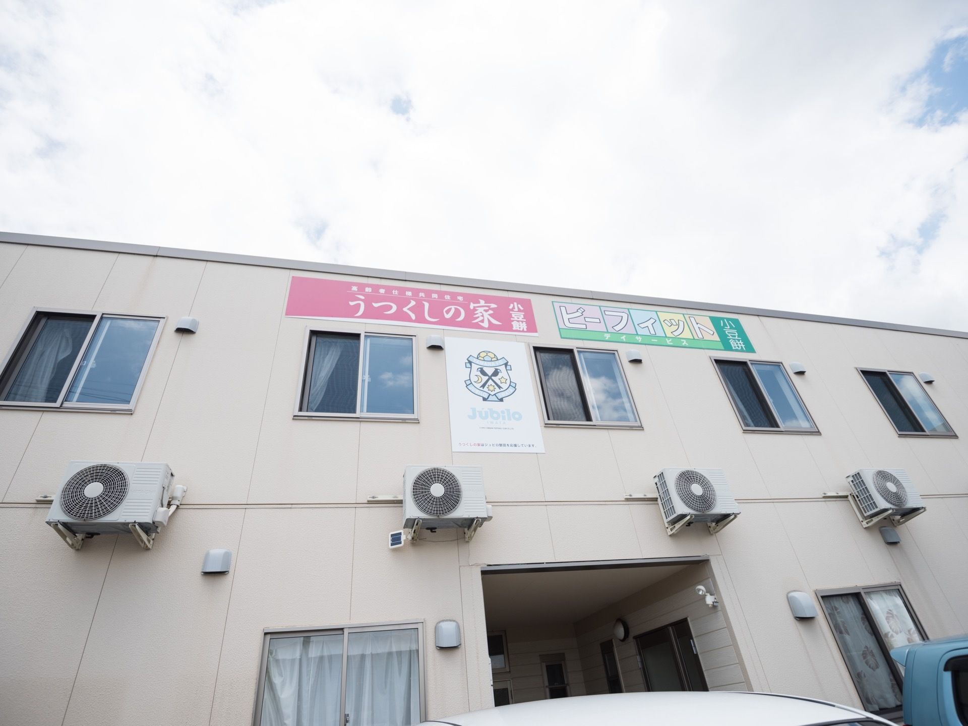 窓が並ぶ２階建ての建物の前に複数の車が停められている。２階の外壁部分に、施設名称を明記した看板が取り付けてある。