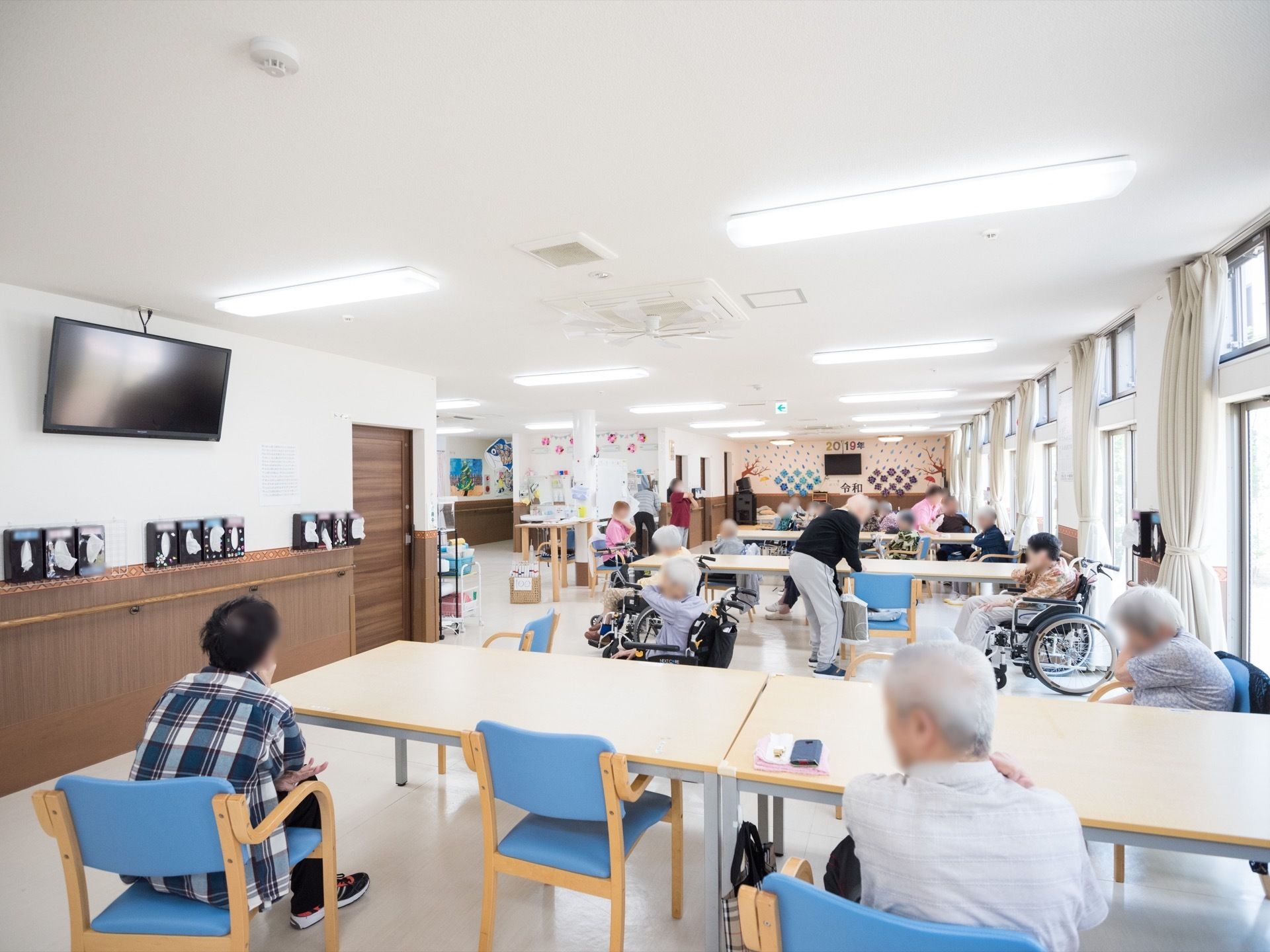 右側の壁面に窓が連続していて、日当たり良好な明るい広間。テーブルに向かって椅子に座っている人やテレビに面している人の姿が見える。
