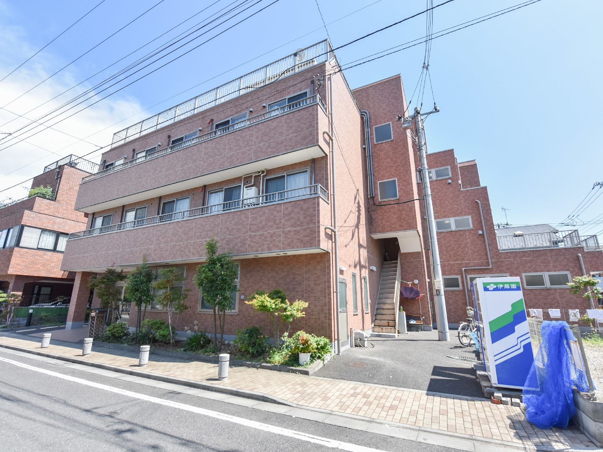 フェンスで敷地が囲まれていて、四階建ての建物である。自動販売機の横に自転車がとめてある。花壇があり、花や木が植えてある。
